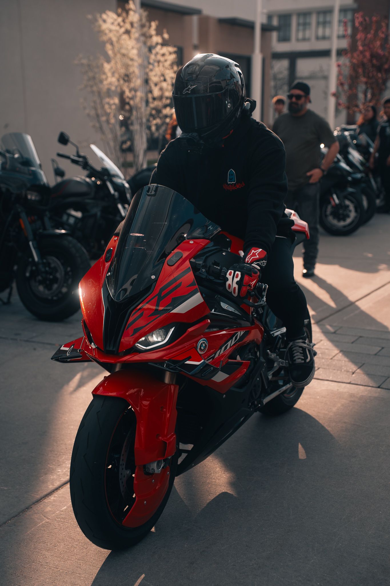 A rider in a black helmet sits on a striking red sport motorcycle surrounded by other bikes in a parking area.