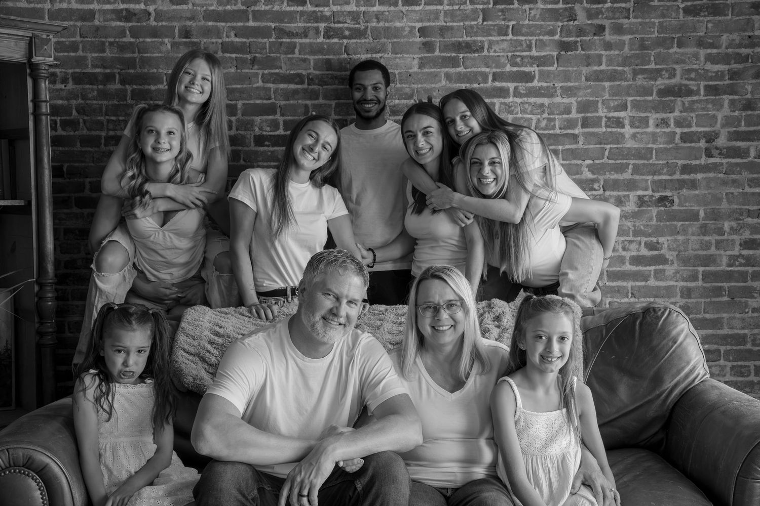 A large happy family poses together in black and white portrait on a couch near a brick wall.