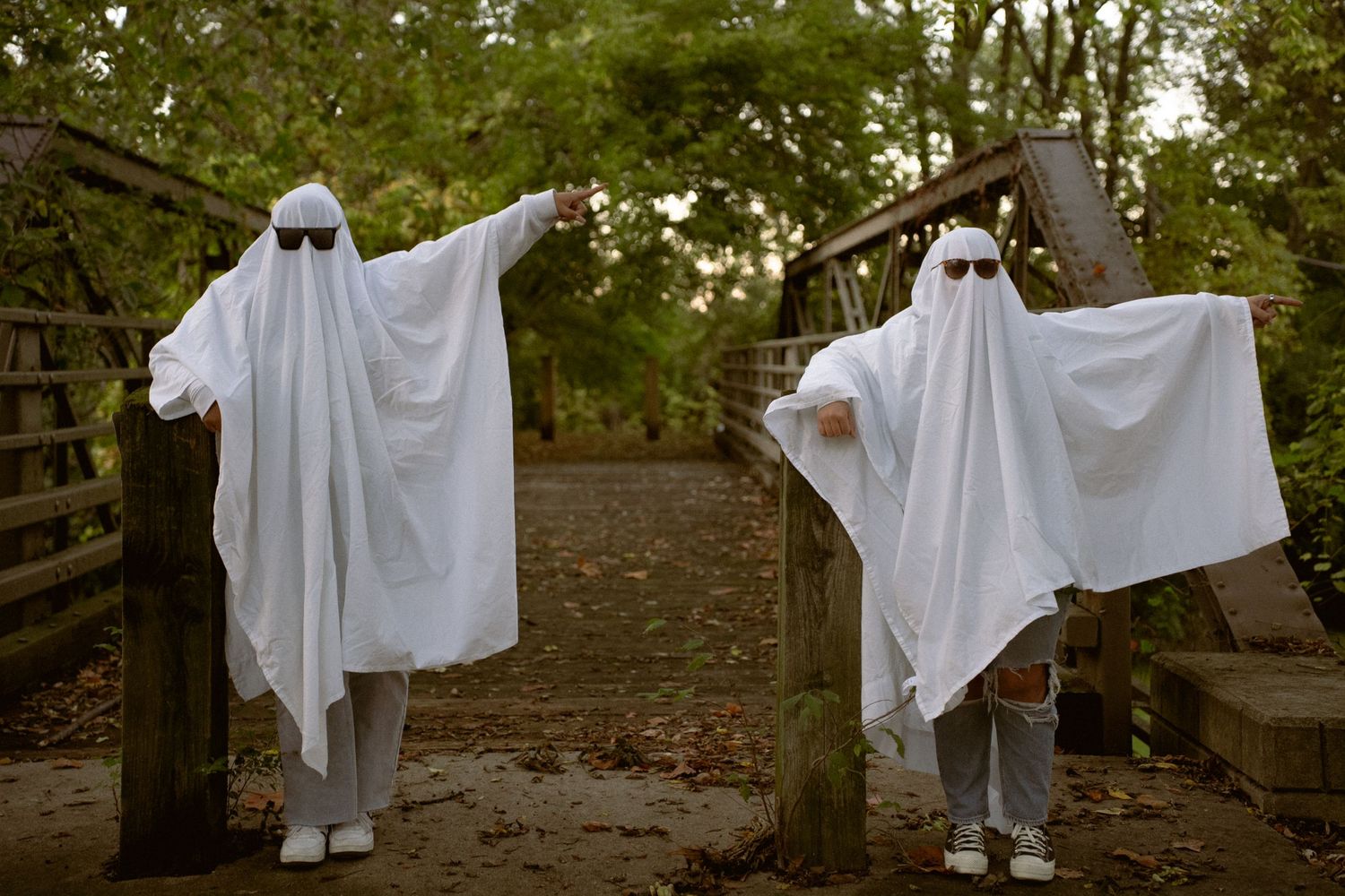 People dressed as playful ghosts in white sheets dance and pose in a wooded area.
