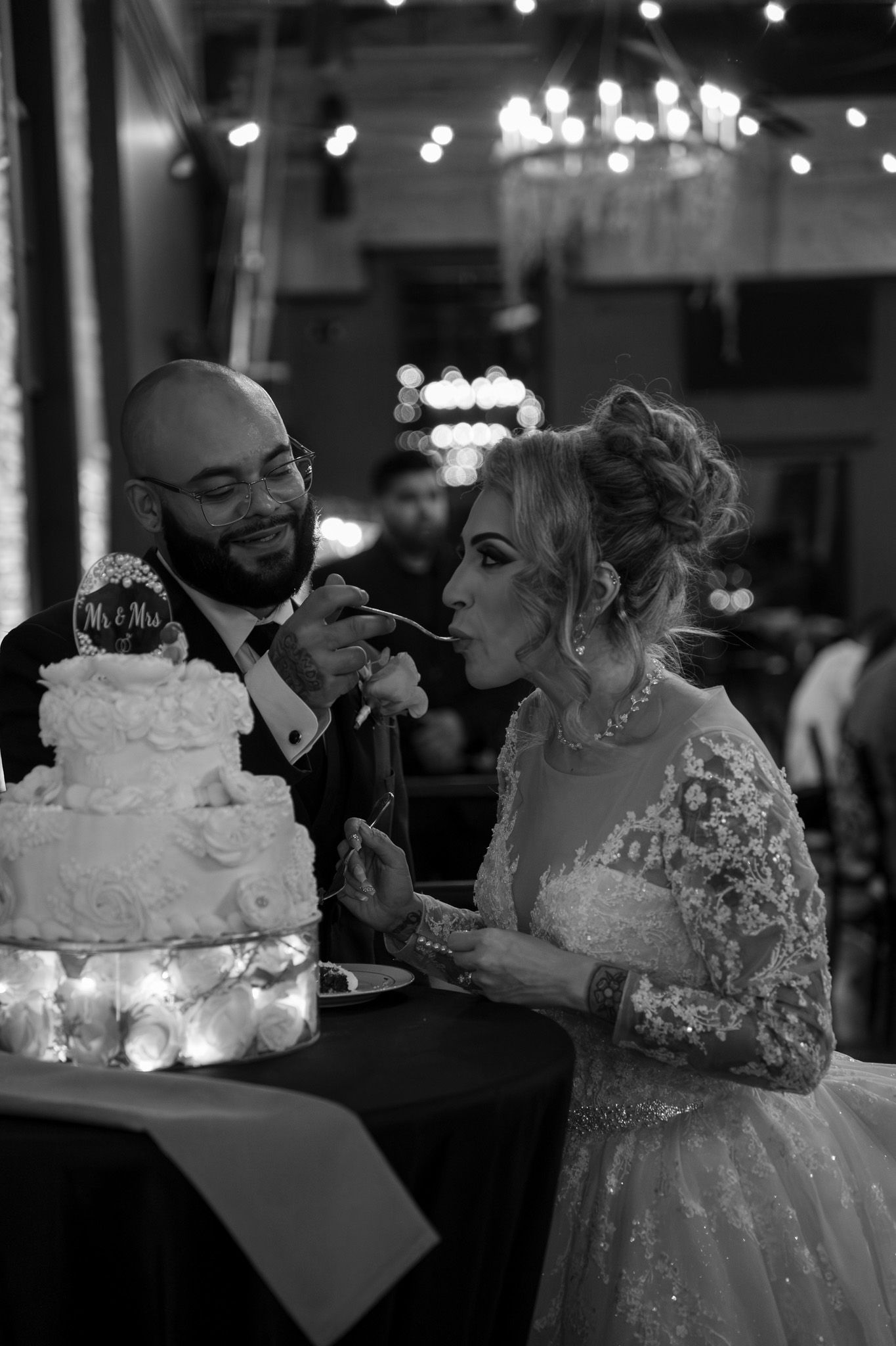 Couple sharing wedding cake at reception with decorative lighting in background.