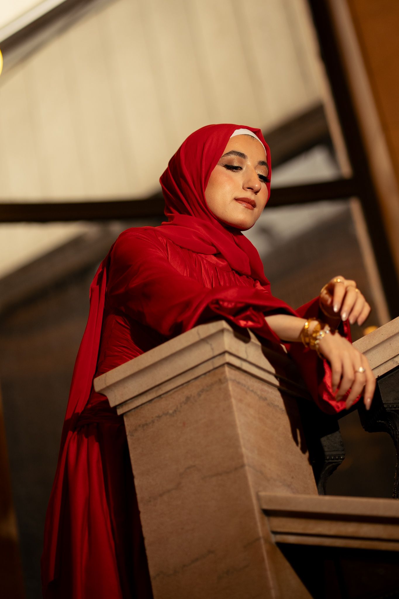 A woman in a red hijab and matching outfit leans gracefully on a stone railing, wearing gold jewelry.