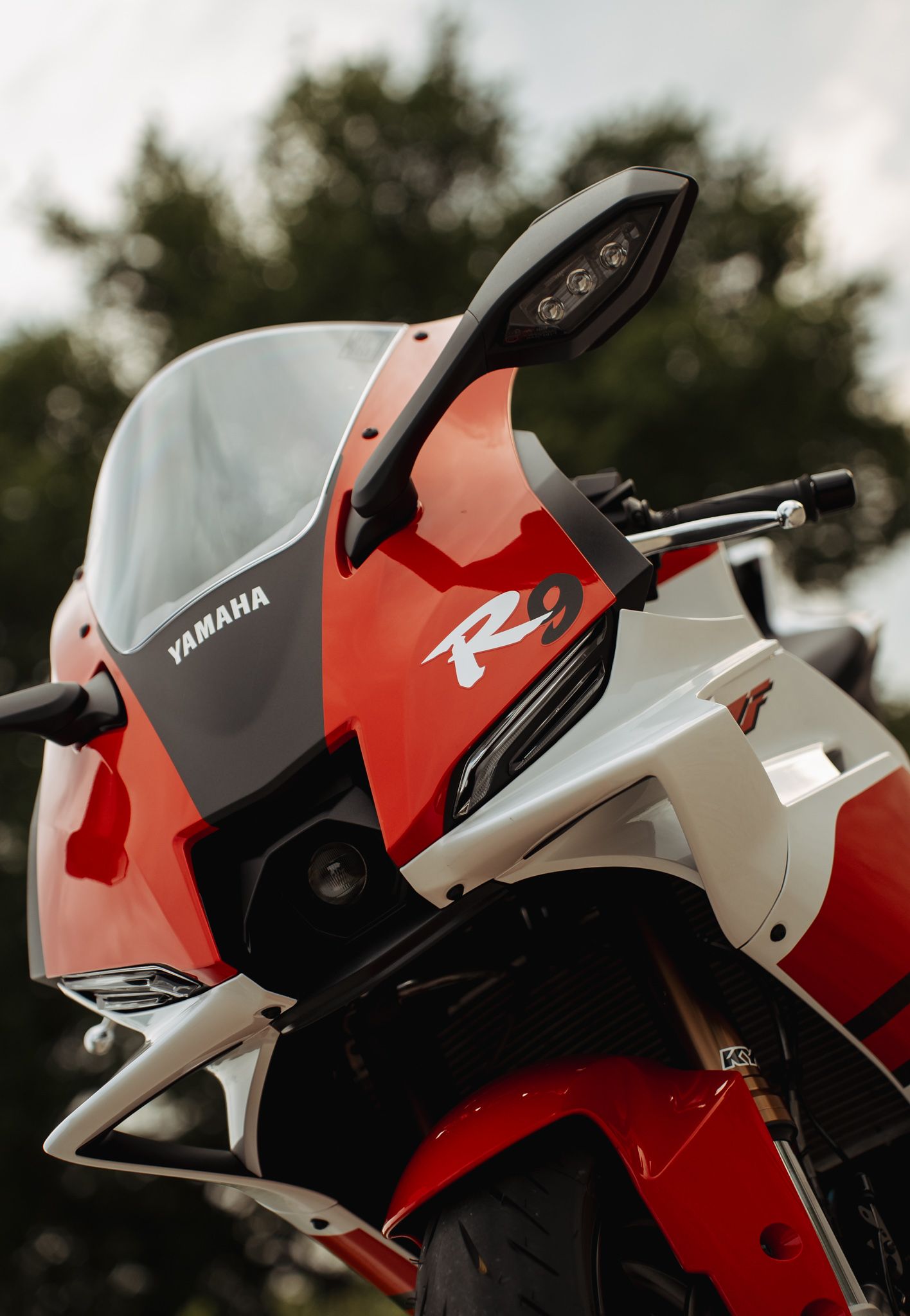 Red and white Yamaha R series sport motorcycle with sleek fairing and windscreen, parked outdoors with trees in background.