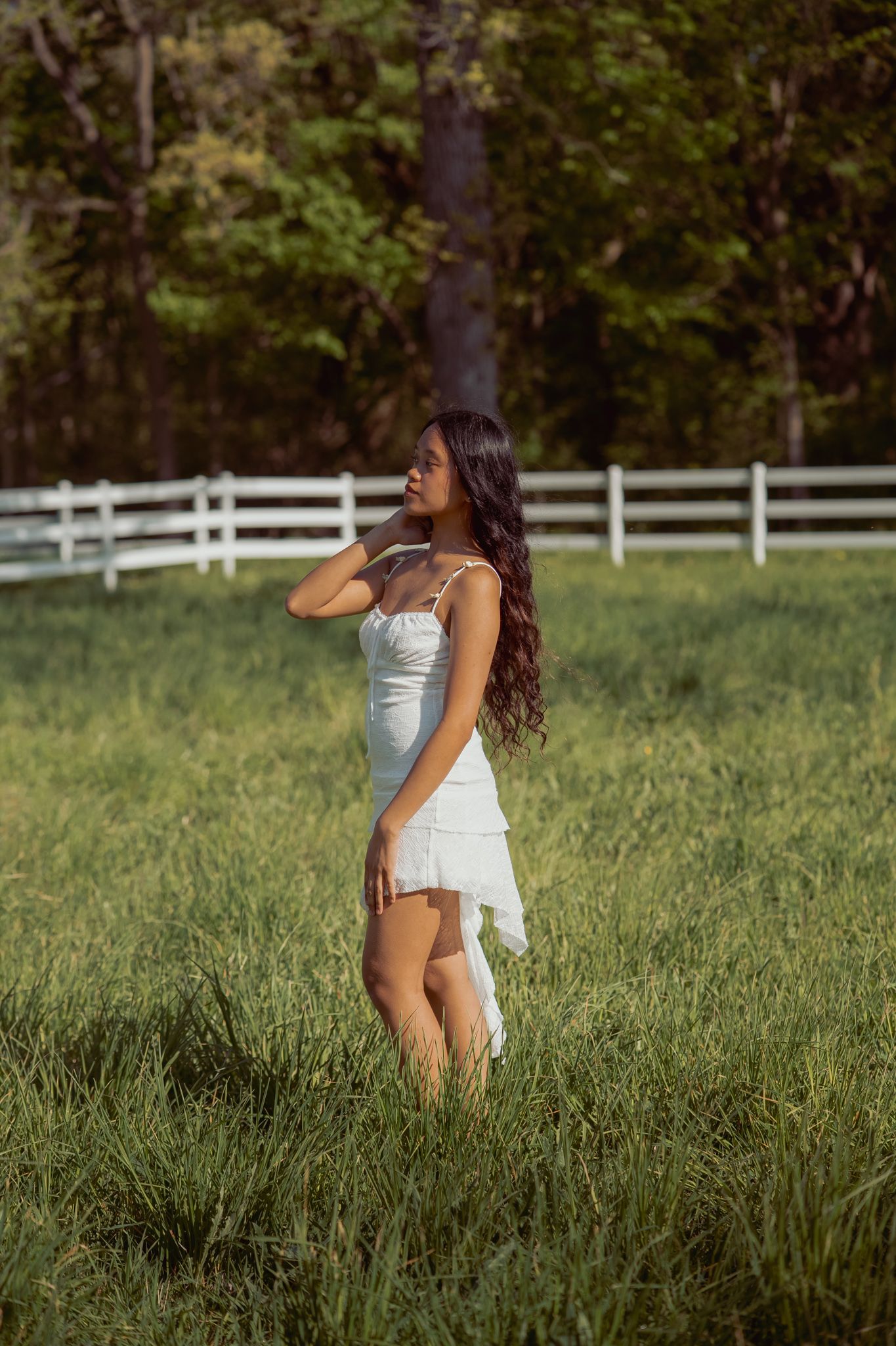 Young woman in white mini dress standing in green field with white fence and trees in background.