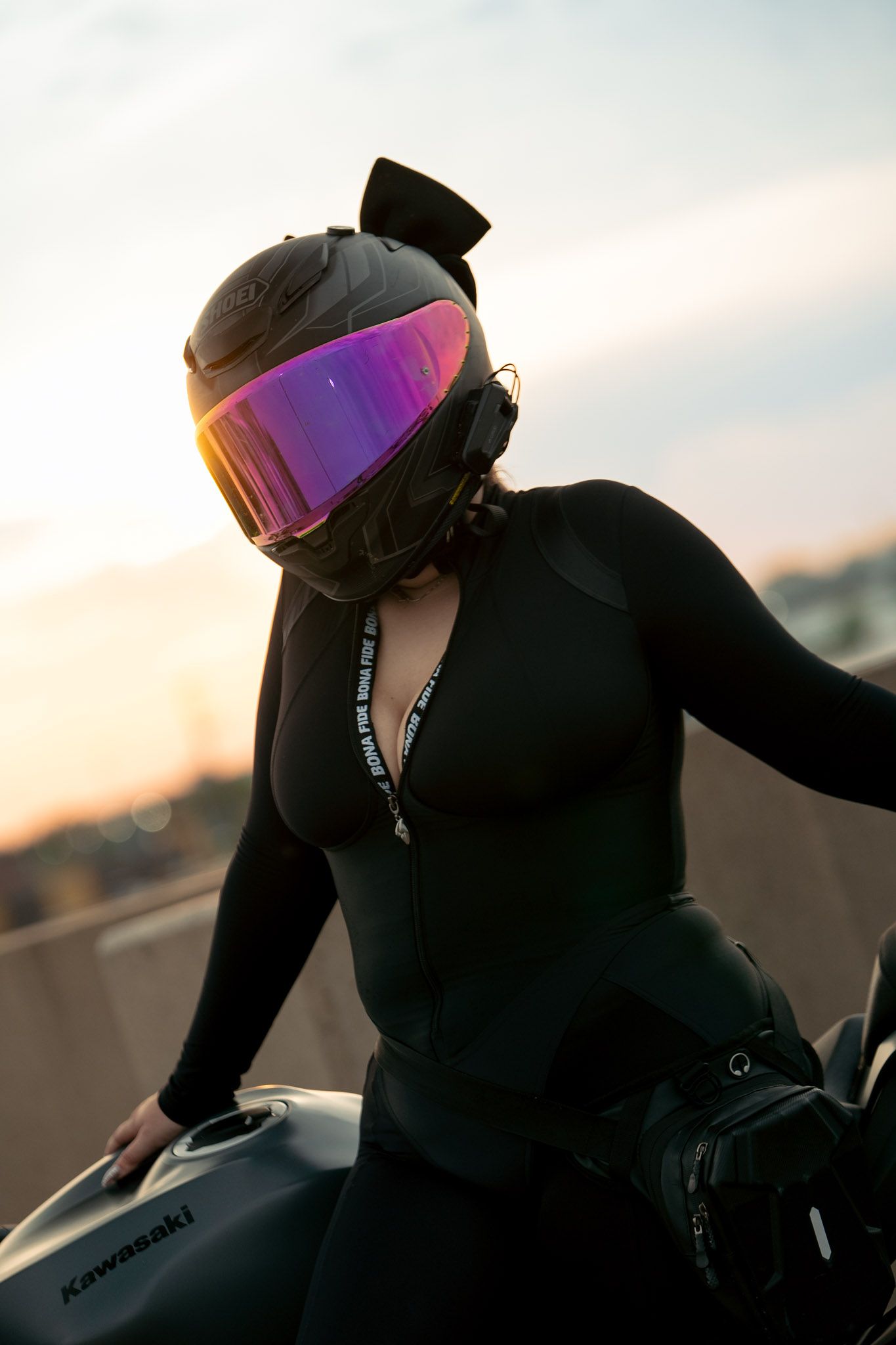 Motorcyclist in black suit and helmet with iridescent visor poses on Kawasaki bike at sunset.