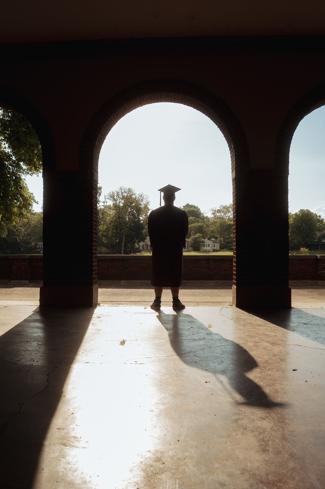 Silhouette of a graduate in cap and gown standing in an arched brick colonnade with sunlit trees in background.