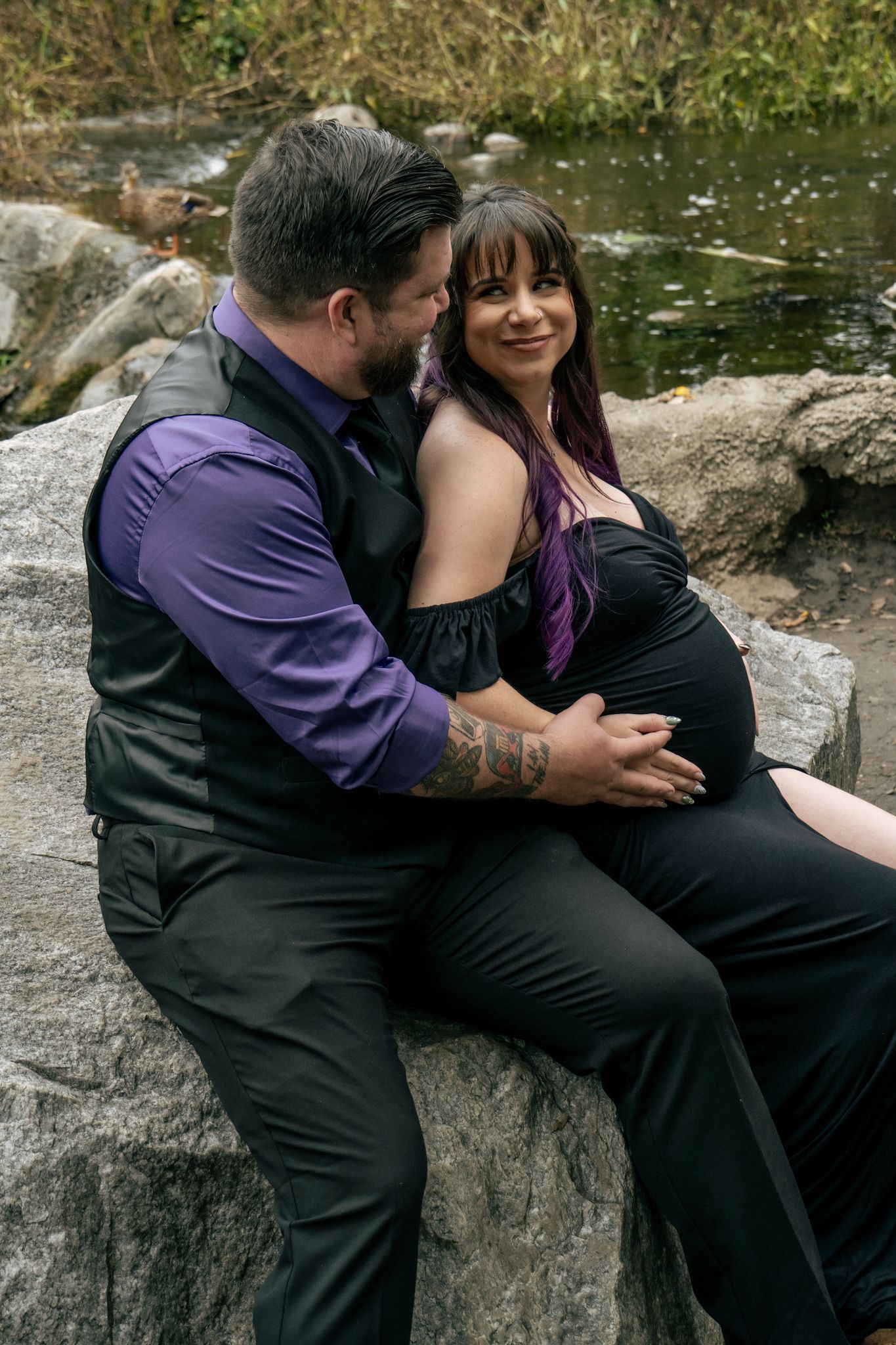 A couple in formal attire shares tender moments while sitting on stone wall by a pond.