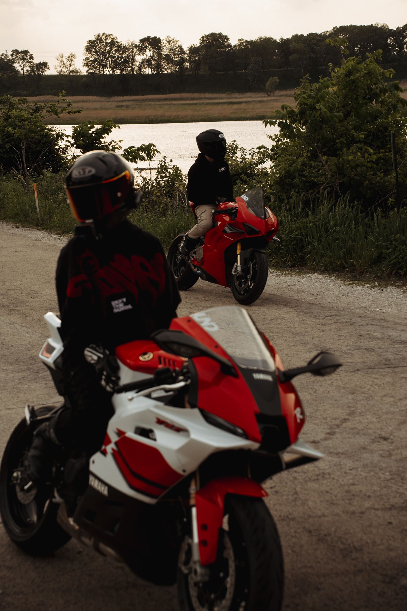 Two motorcyclists on red sport bikes pause on a gravel path near a scenic lake surrounded by lush greenery at dusk.