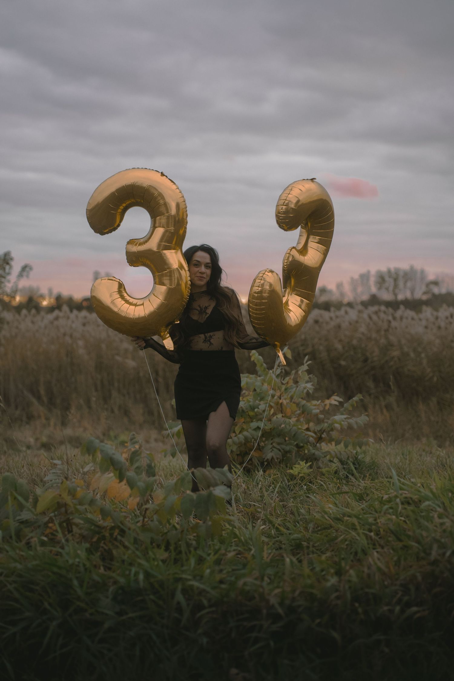 A person poses with giant gold number 33 balloons in a moody outdoor setting.