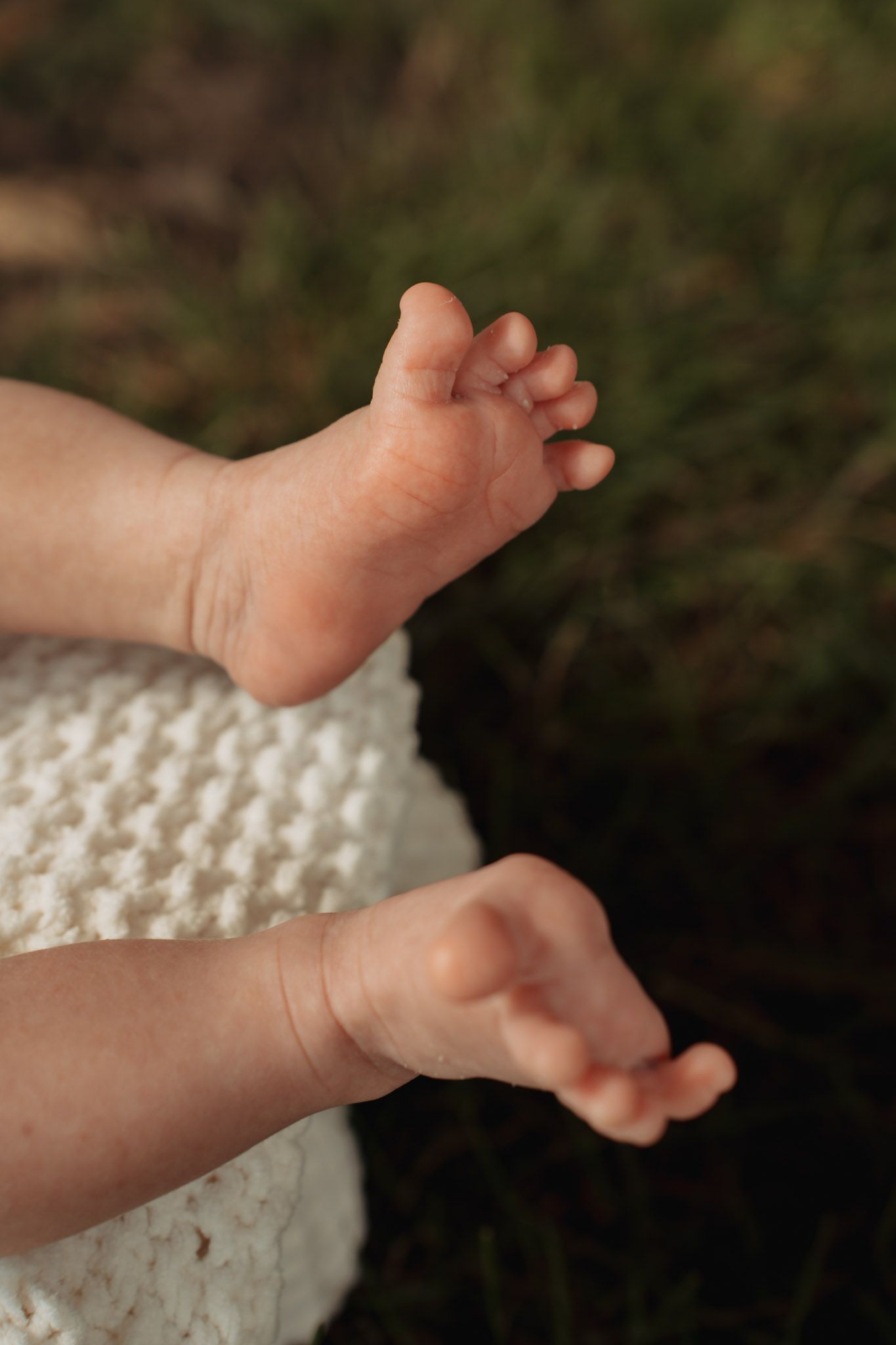 Close-up of tiny newborn baby feet and hands resting on a white knit blanket outdoors.