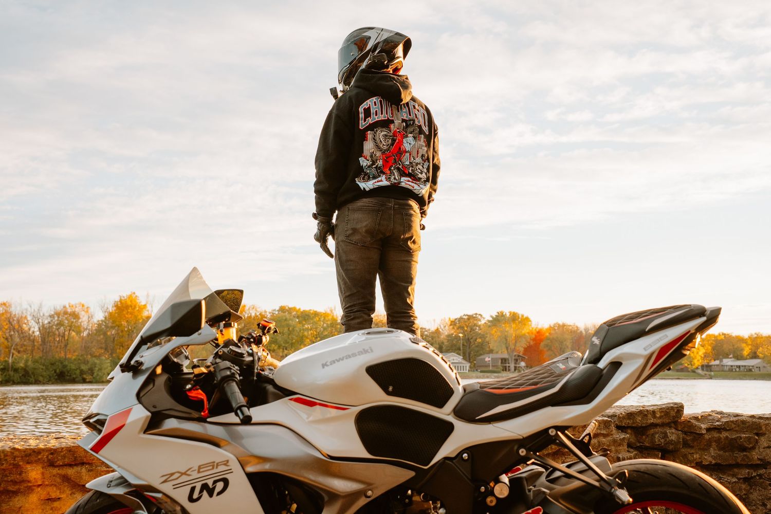 A white motorcycle is parked against a scenic autumn landscape with trees and water in the background.