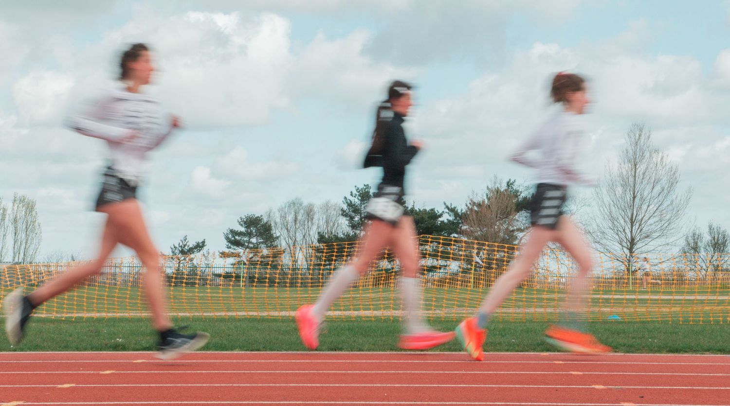 Three runners racing on a red athletic track outdoors, captured with motion blur on a cloudy day.
