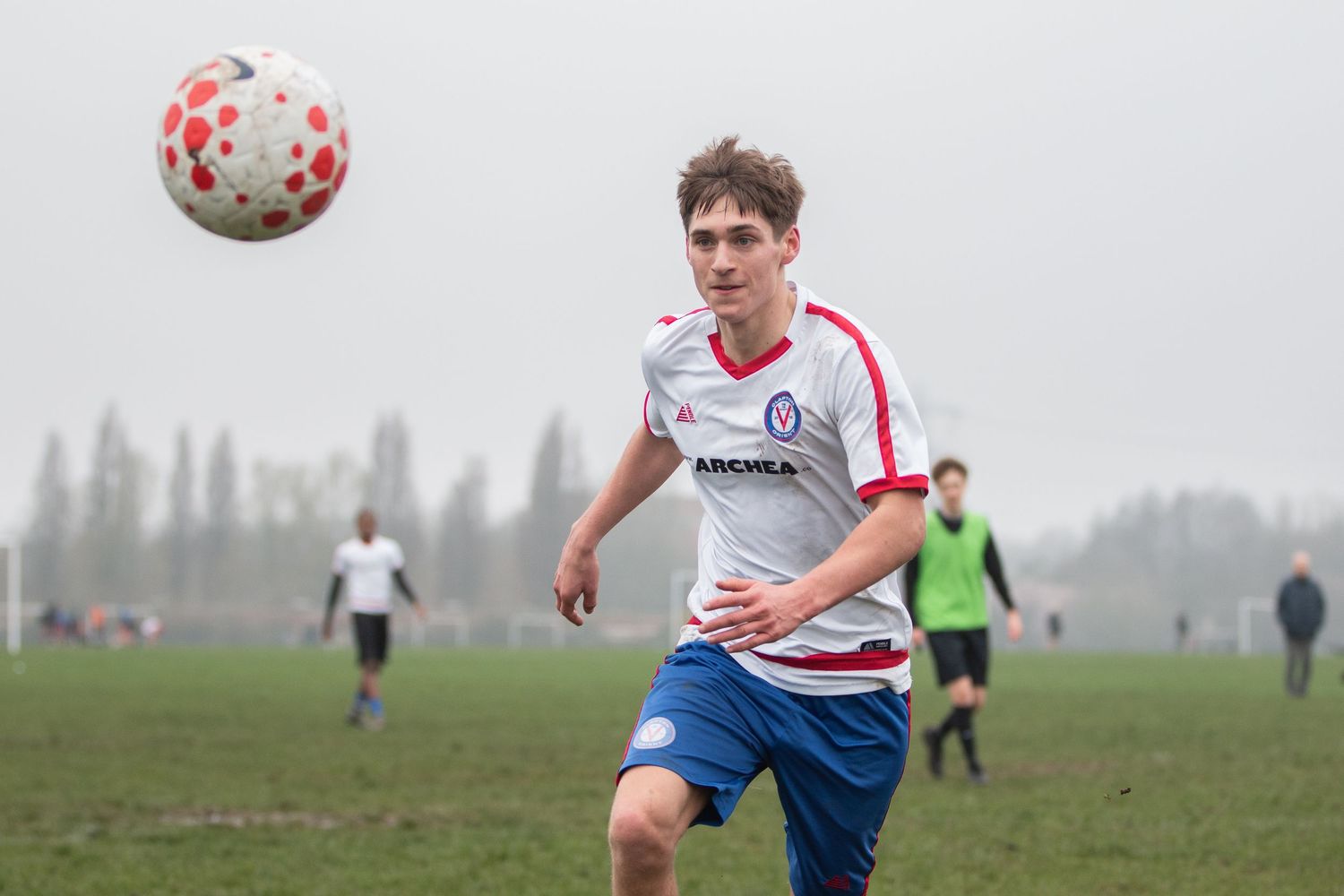 Soccer player in white and red jersey chases after a ball during a match on a muddy field.