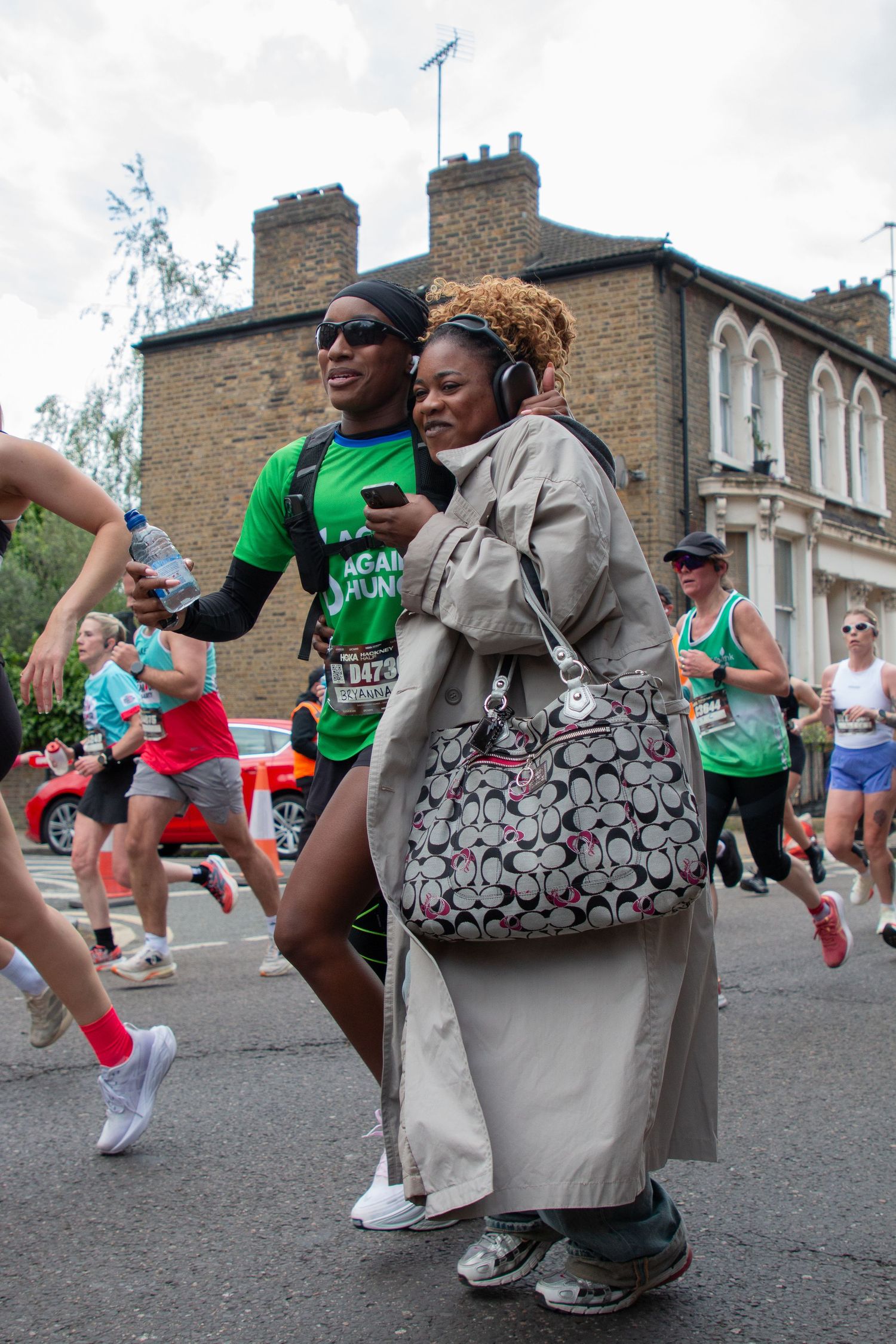 Two people pose together during a marathon race on a city street, with runners passing by in the background.