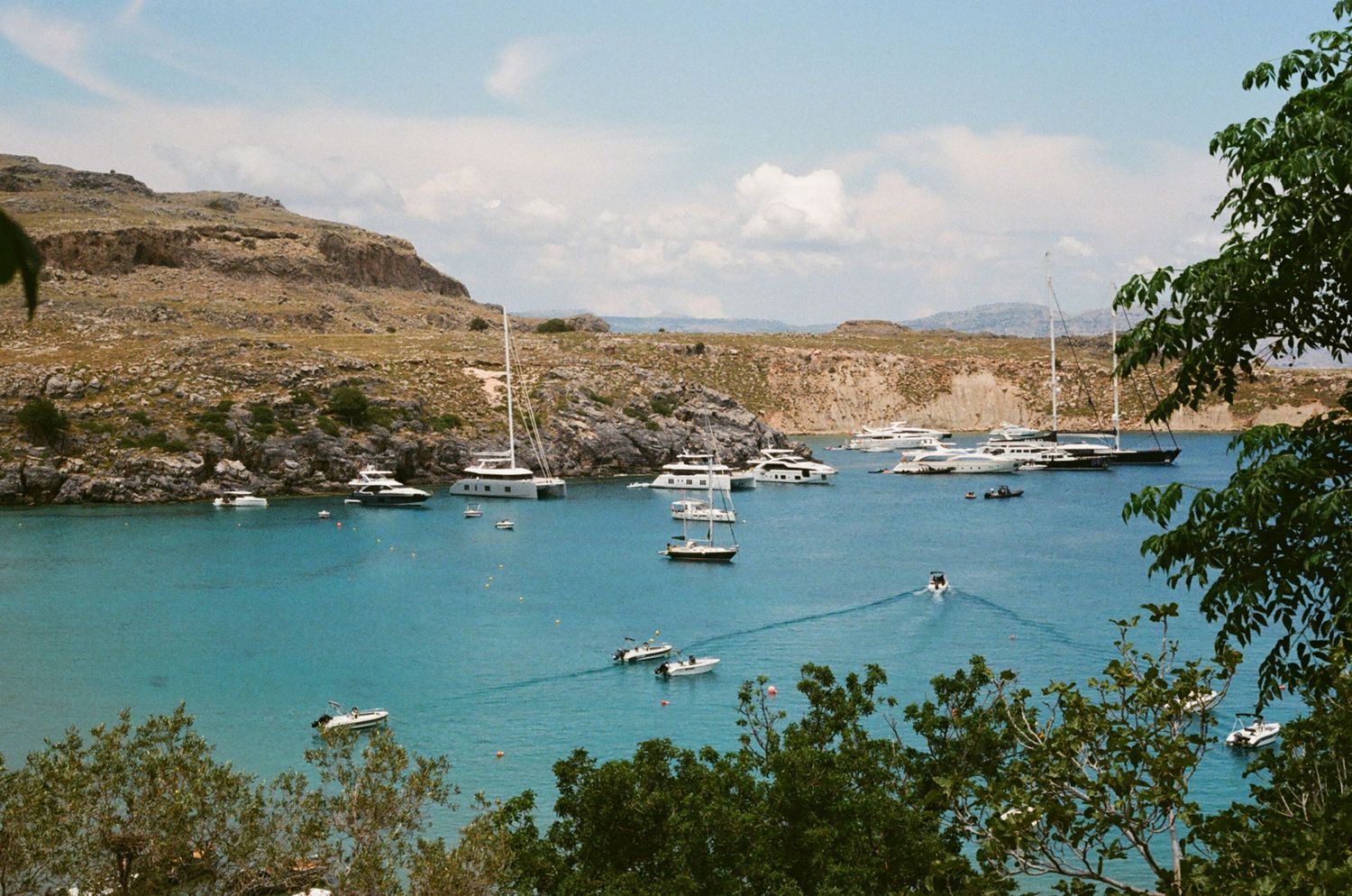 Turquoise harbor filled with anchored boats surrounded by rocky cliffs and hills.