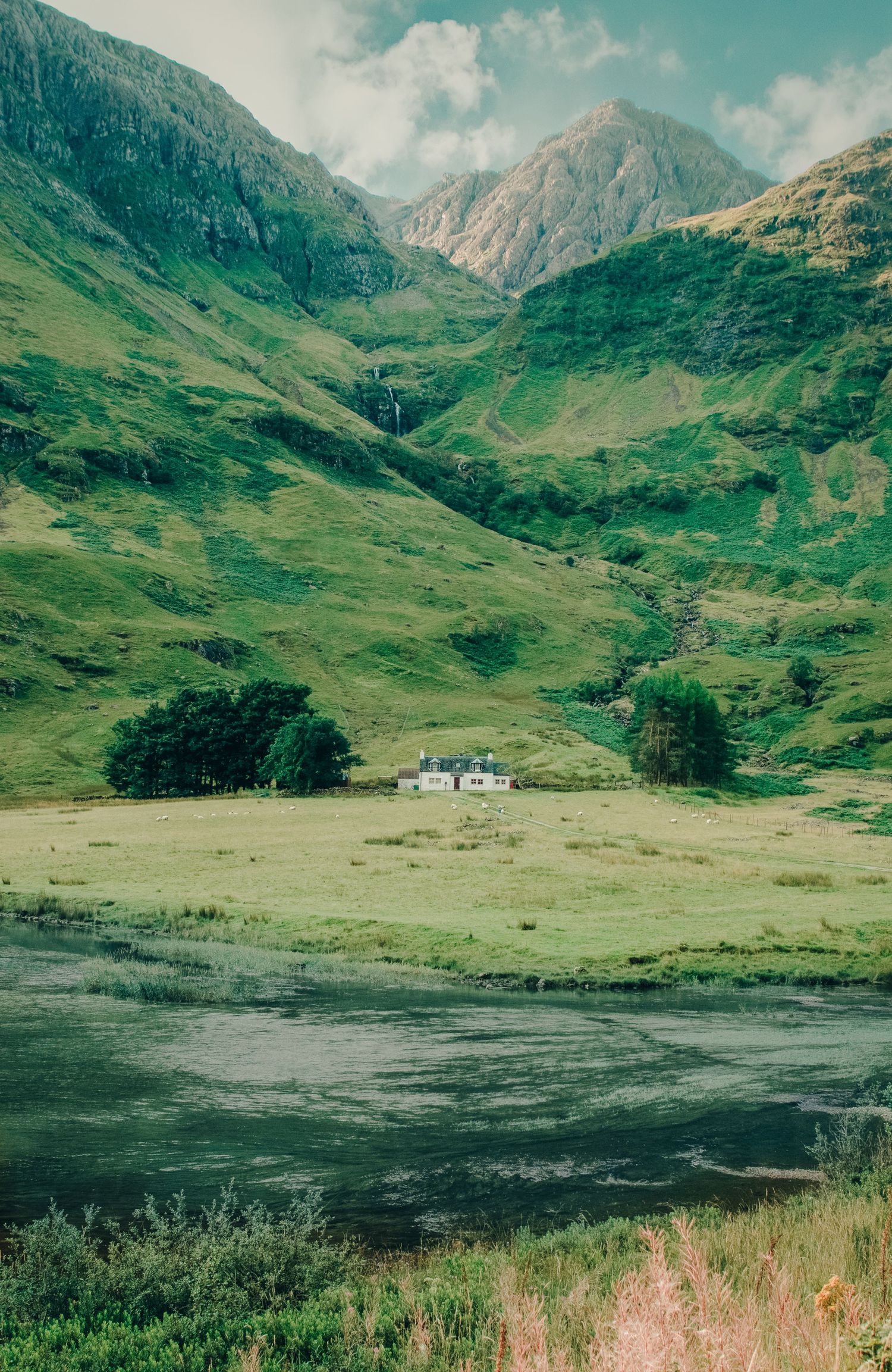 Lush green Scottish Highland valley with a white cottage nestled between rolling mountains and a calm reflective lake.
