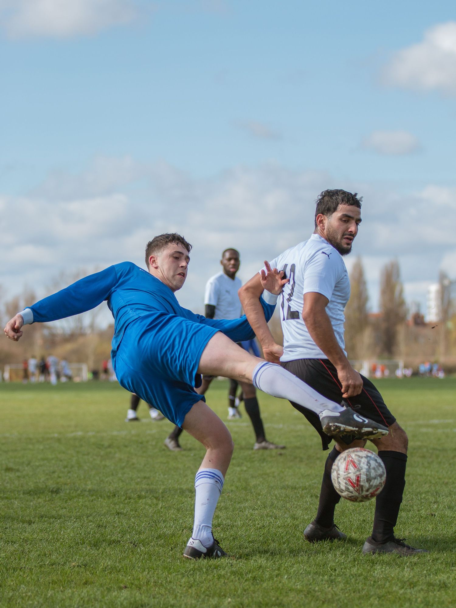 Two soccer players compete for the ball during an amateur match on a grass field under blue skies.