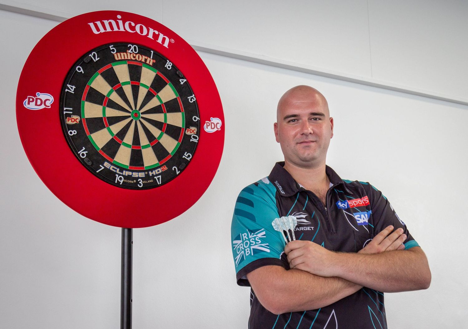 Professional darts player, Rob Cross, stands confidently with arms crossed in front of a red-framed dartboard.