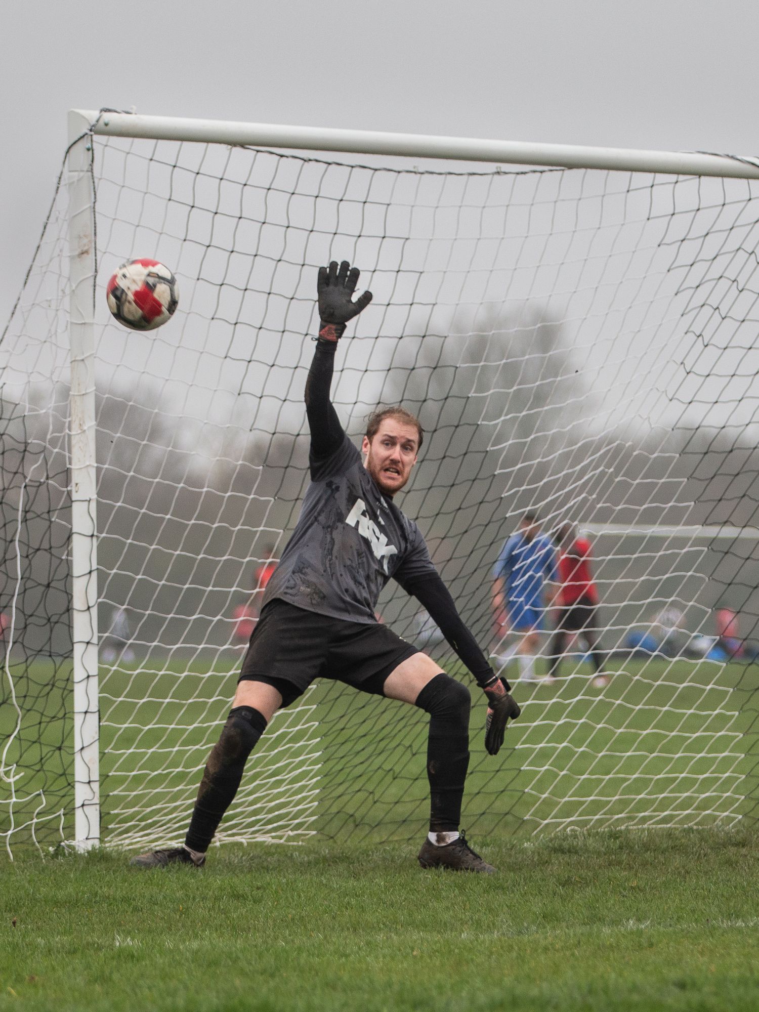Soccer goalkeeper in black uniform stretches to make a save during a foggy match on a grass field.