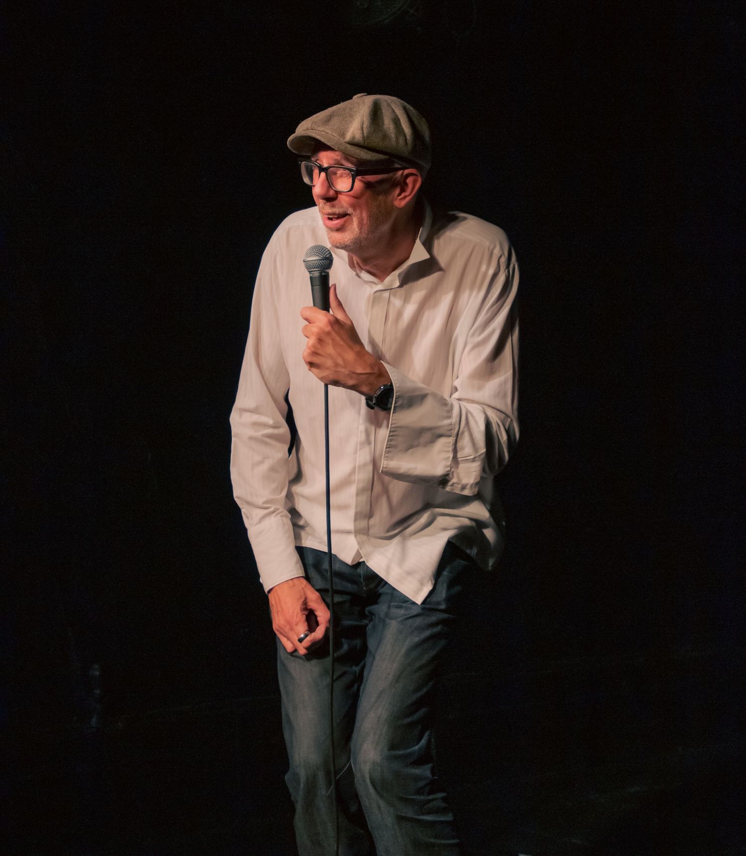 Performer in casual attire delivers comedy routine on dimly lit stage.