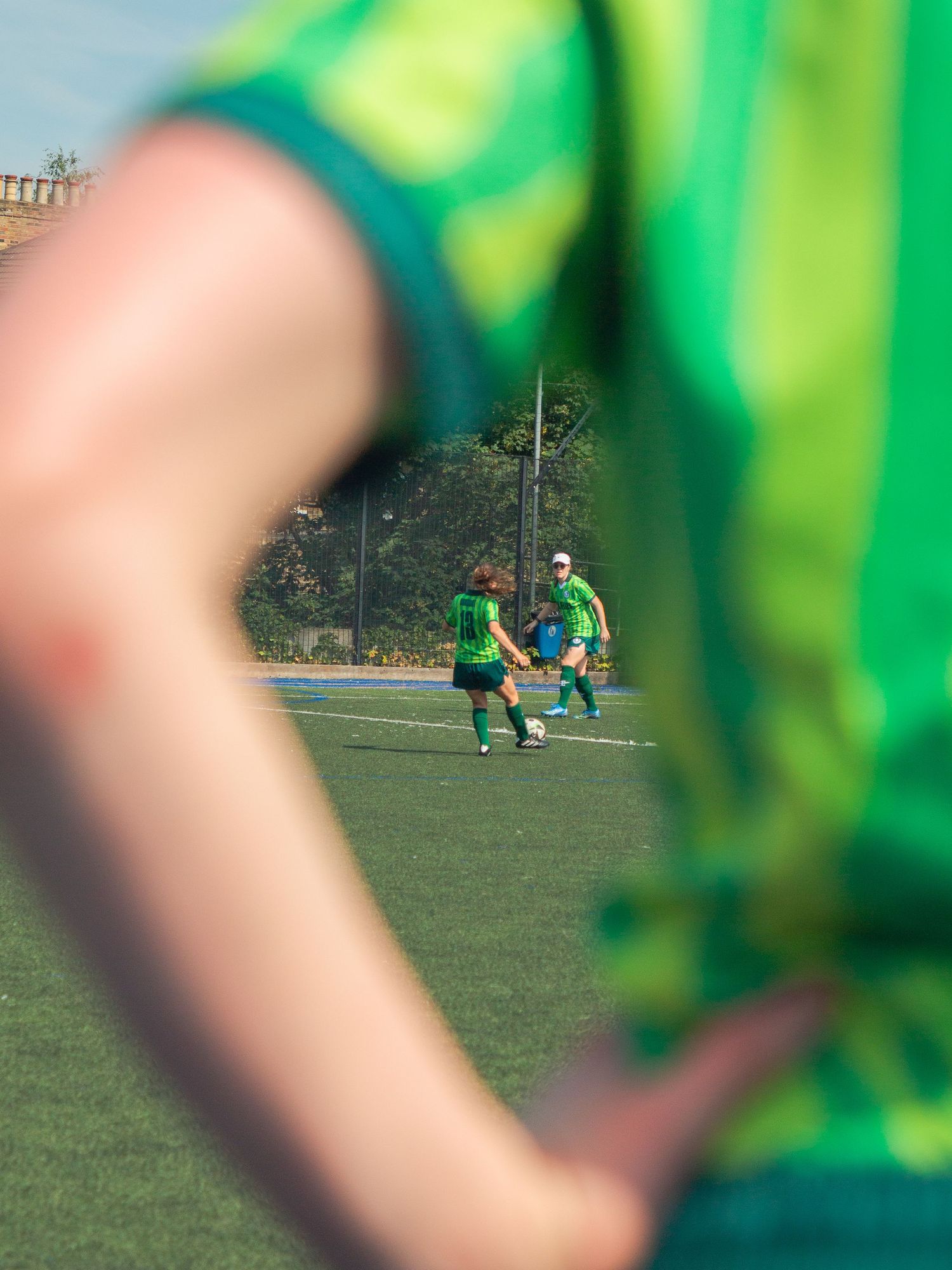 Players in green jerseys compete on a soccer field, viewed through a blurred foreground player during a sunny match.