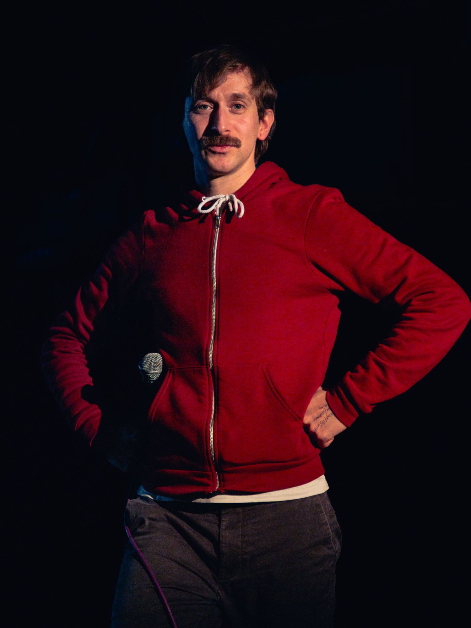 Man with mustache wearing red zip-up hoodie stands confidently with hands on hips against dark background.