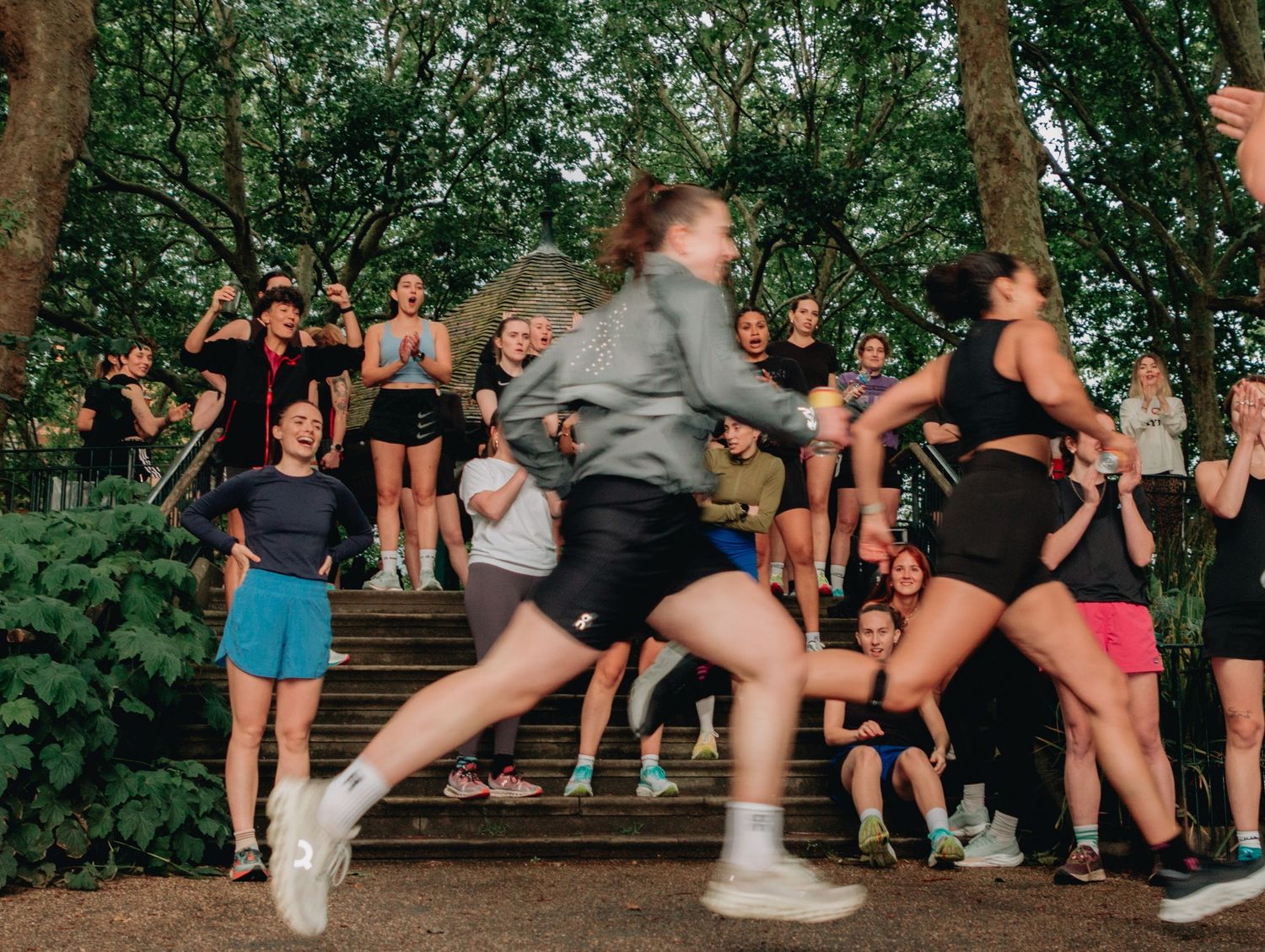 Women running and cheering on outdoor stone steps surrounded by trees during a group fitness event.