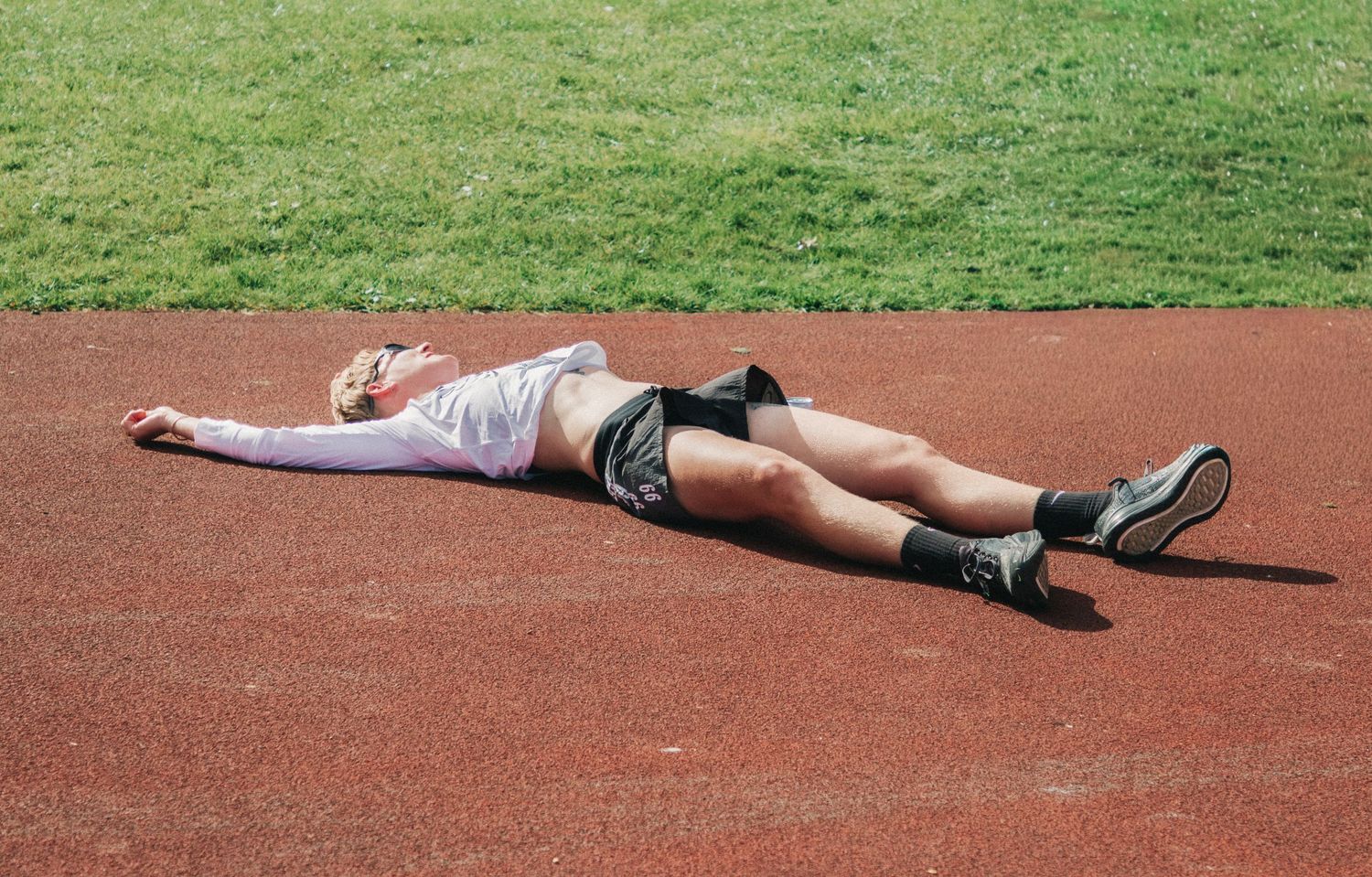 Exhausted athlete lying flat on red running track after race, wearing shorts and sneakers, resting on sunny day.