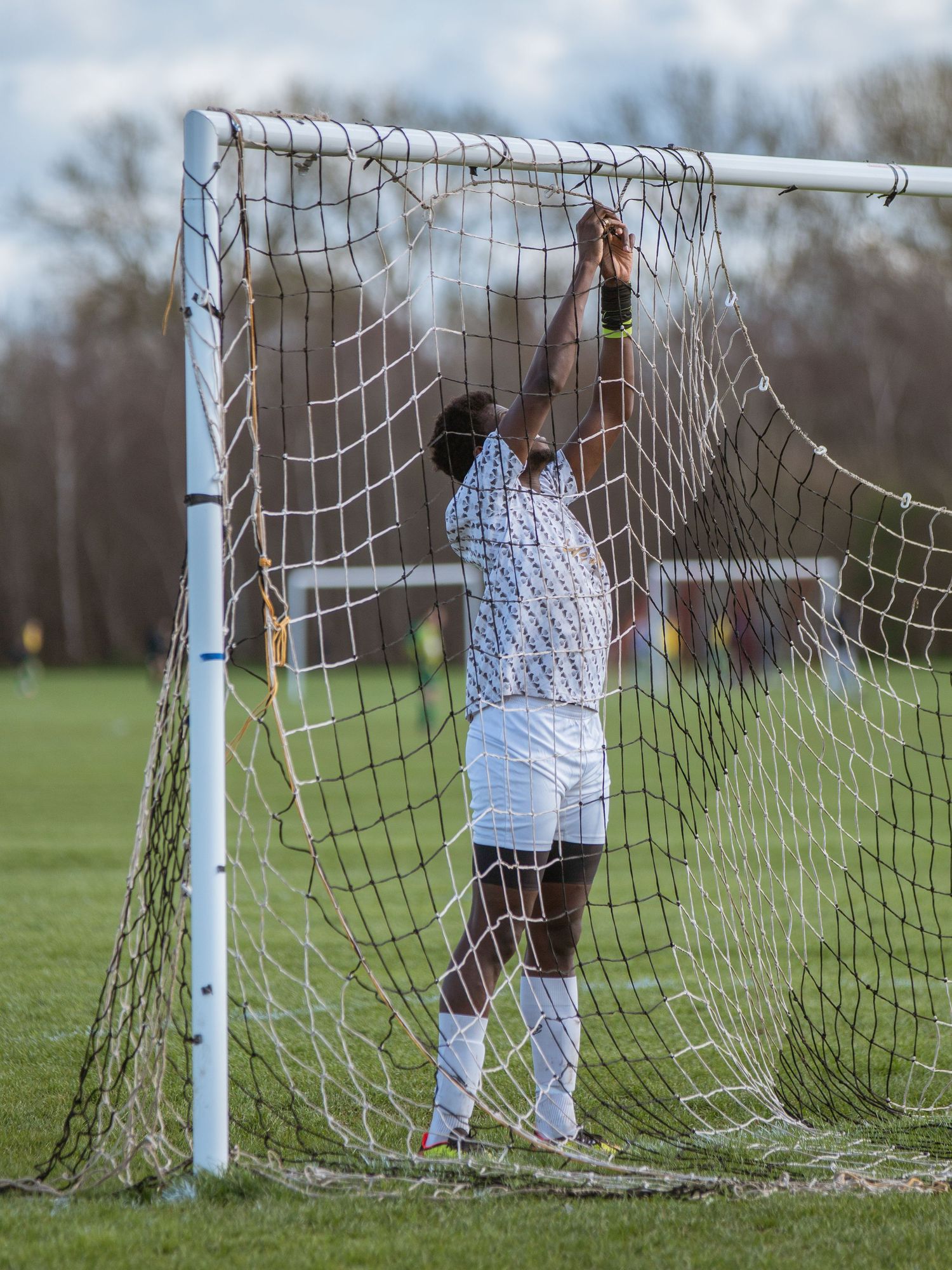 A soccer player in white uniform reaches up to adjust the goal net during a match on a grass field.