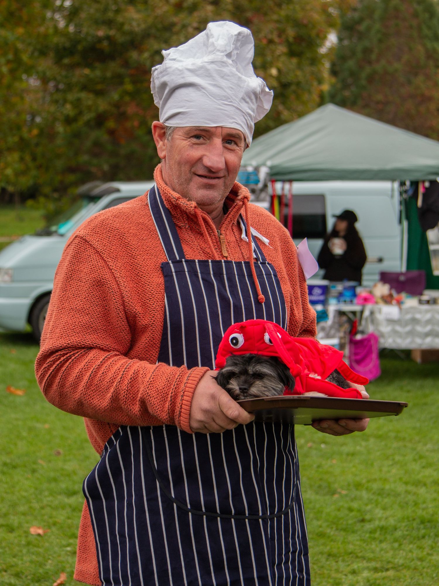 A person wearing a striped apron and orange sweater serves his dog dressed as a lobster for a community dog competition event.