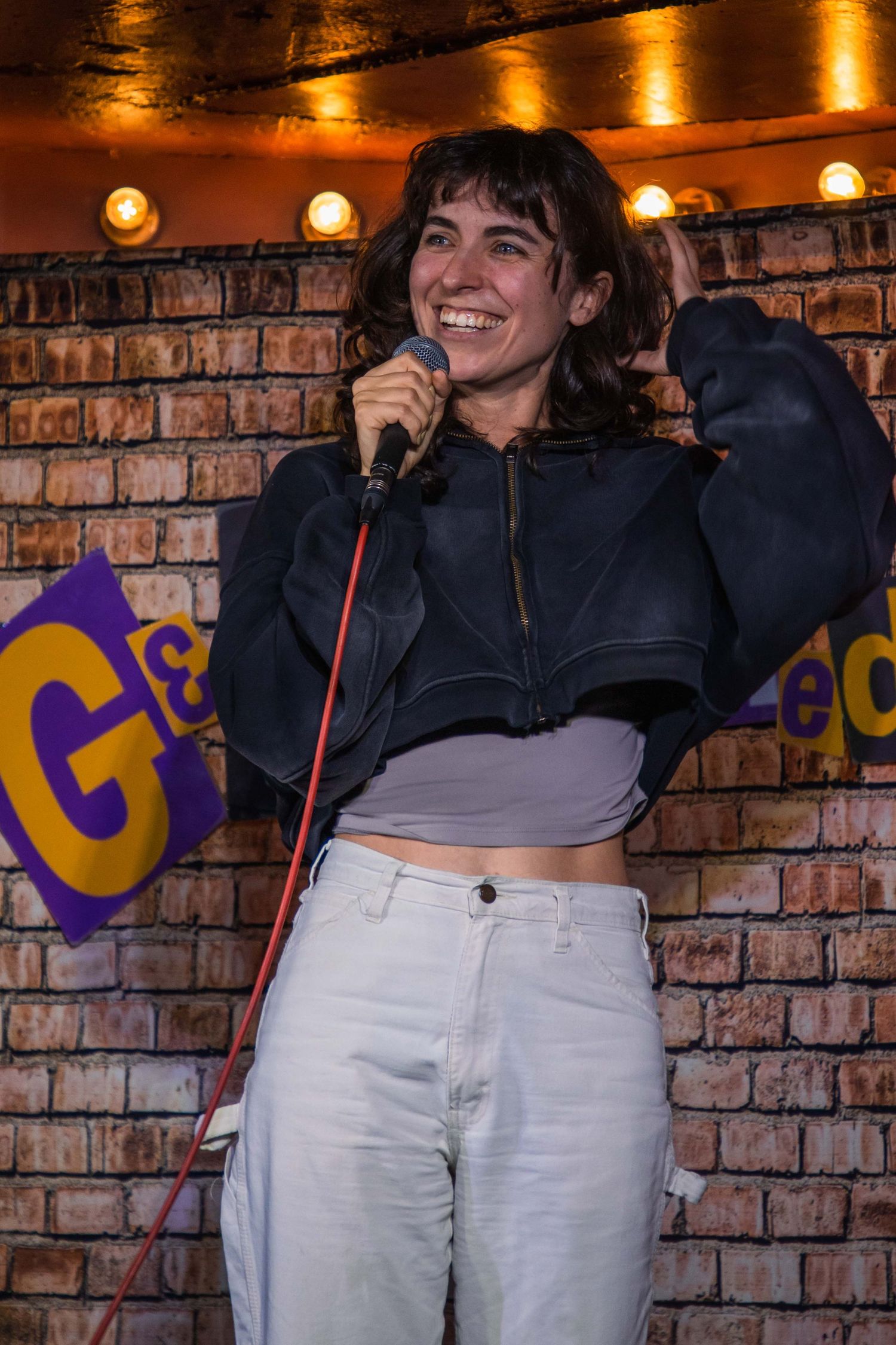 A performer in a black jacket and white pants sings on stage at a comedy club with brick walls and stage lighting.