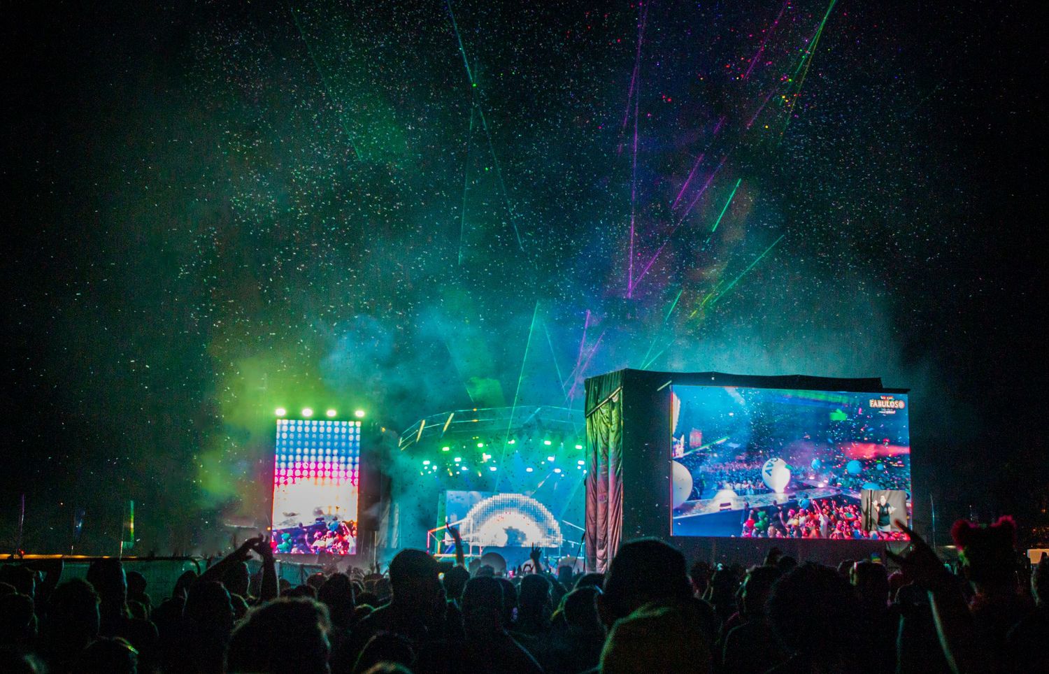 Large outdoor music festival crowd silhouetted against colorful stage lights and visual effects.