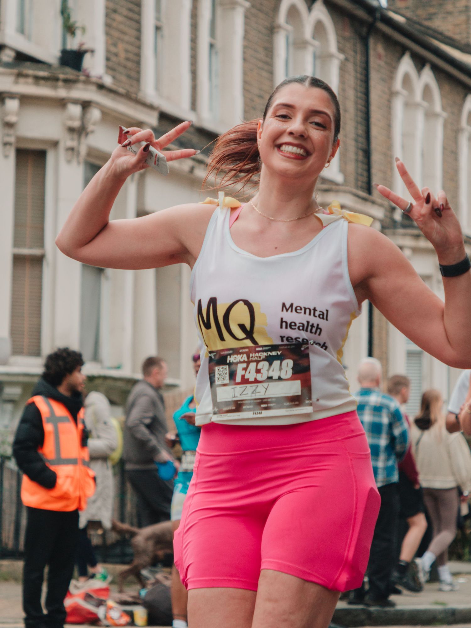 Smiling woman in MQ Mental Health vest and pink shorts making peace signs at a charity marathon race.