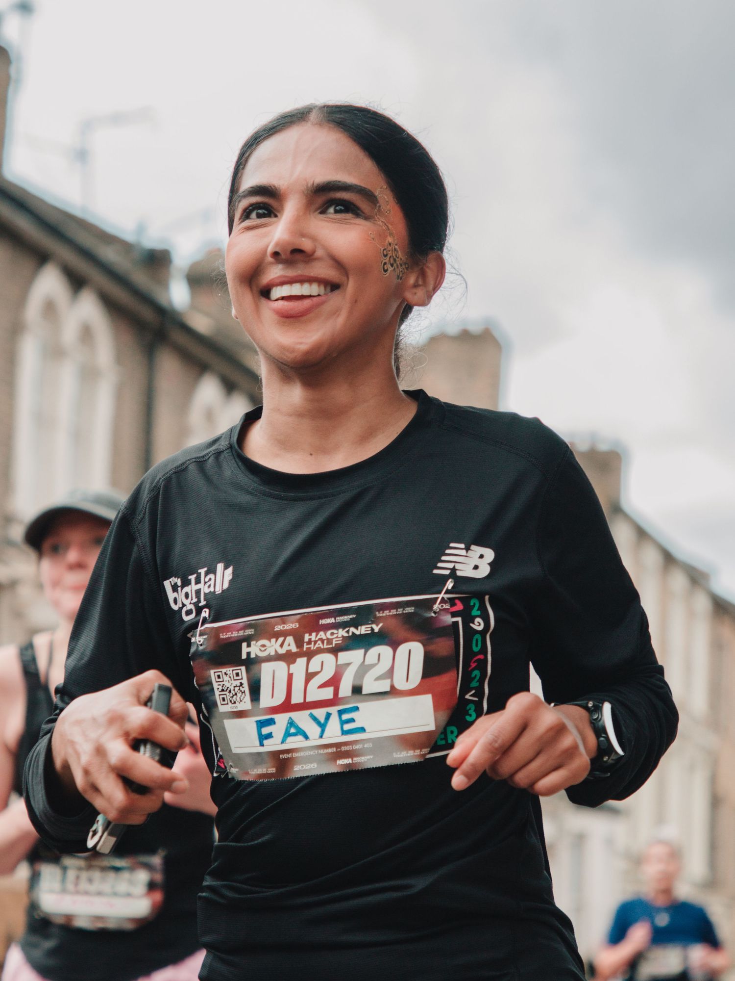 Smiling female runner named Faye wearing bib number D12720 at the HOKA Hackney Half Marathon in a black New Balance top.