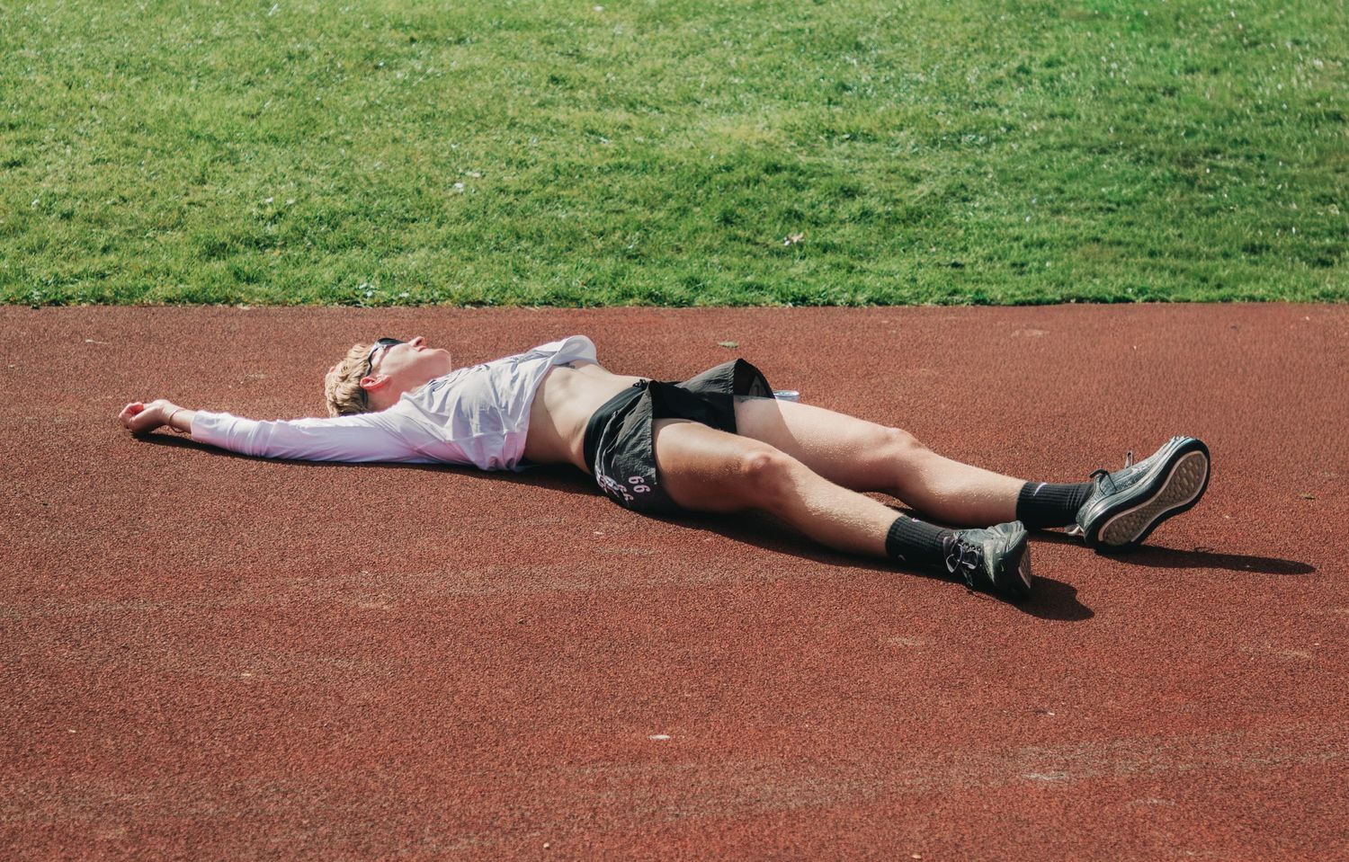 Exhausted athlete lying flat on a red running track after a workout, wearing shorts and sneakers.