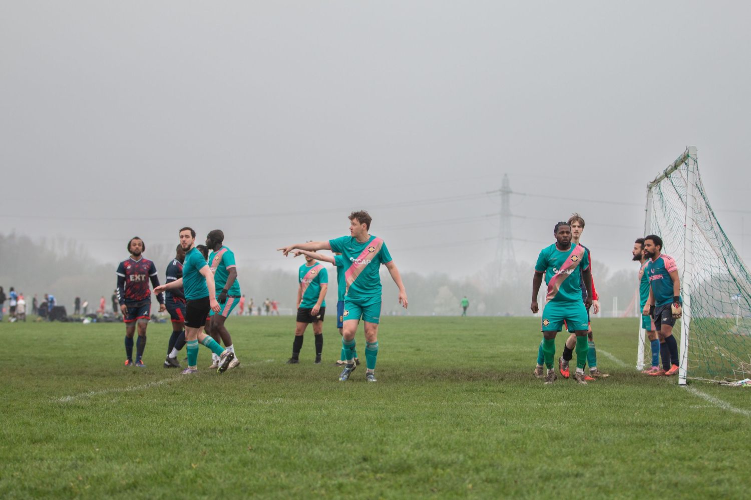Soccer players in teal jerseys compete during a foggy match on a grass field.
