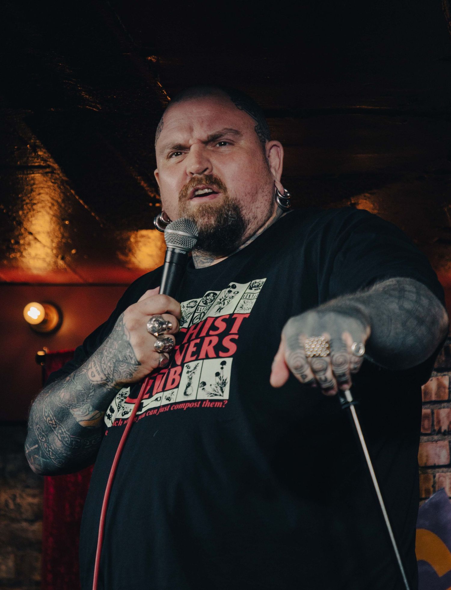 Tattooed comedian gesturing dramatically during performance on dimly lit stage.