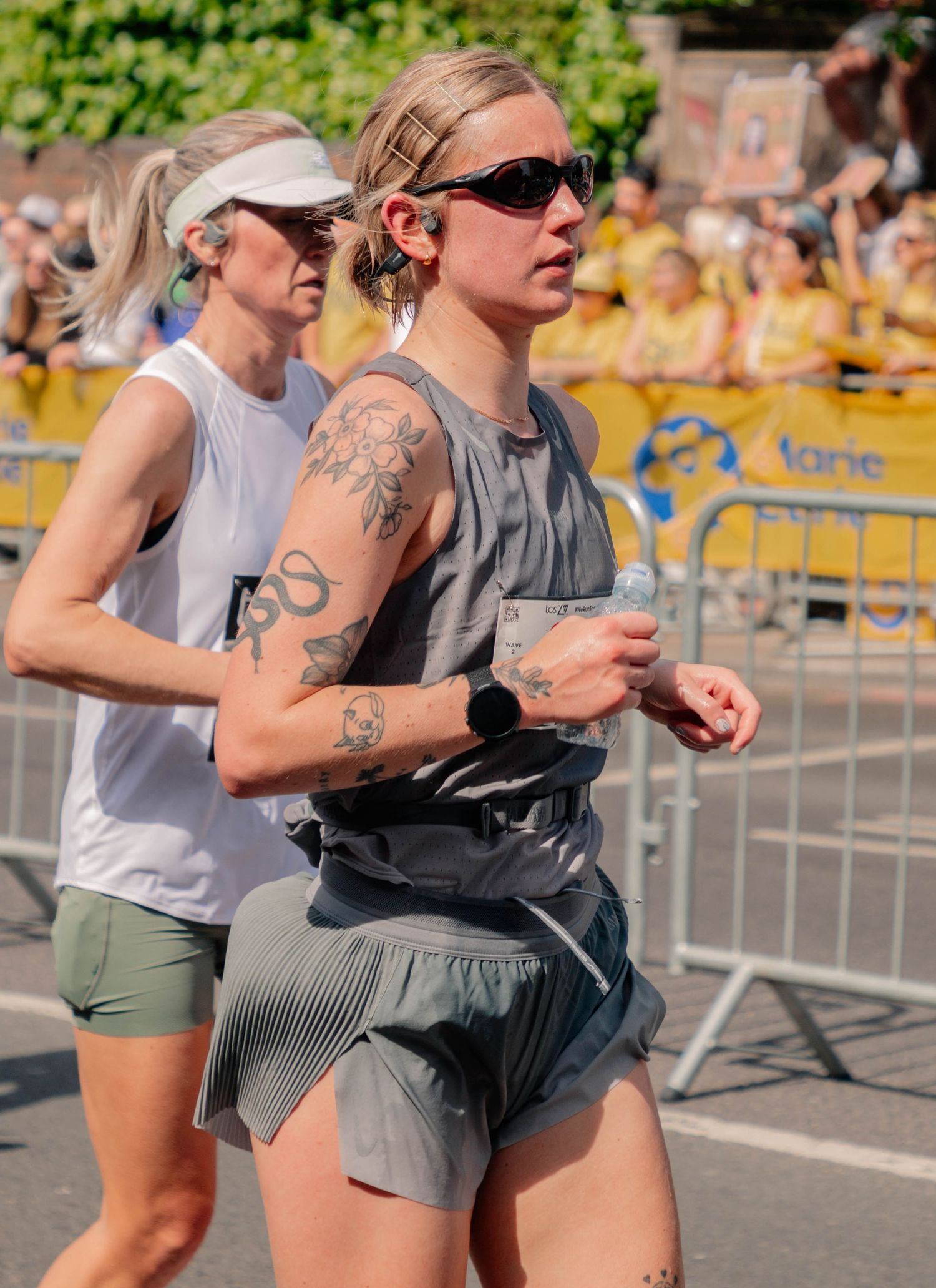 Two female runners competing in an outdoor race, one with tattoos and sunglasses holding a water bottle.