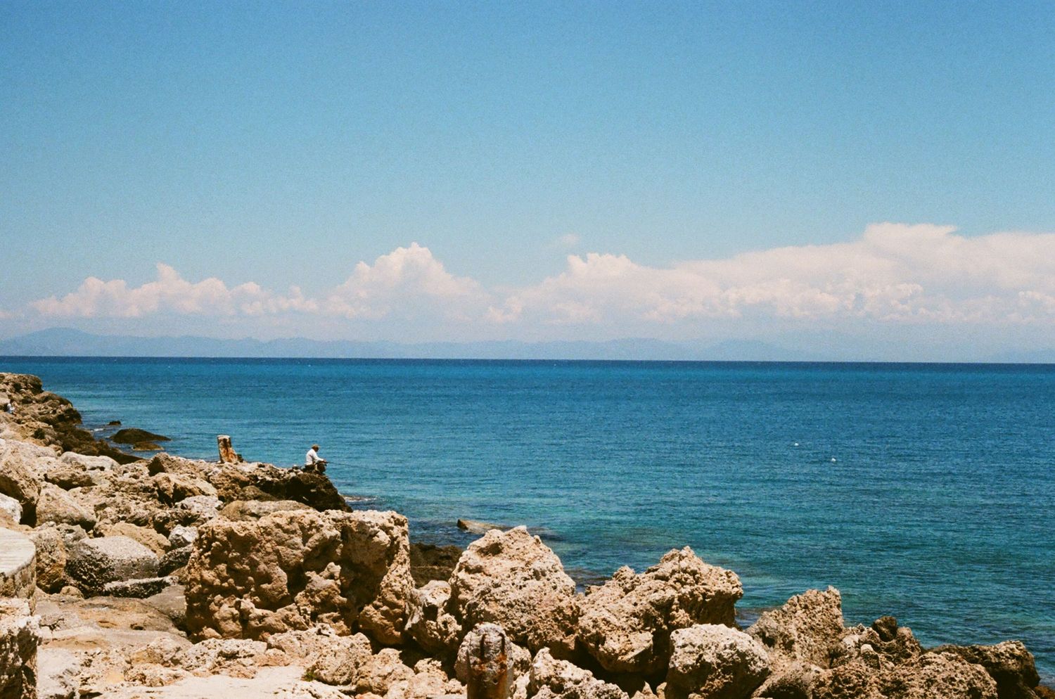 Rocky coastline overlooking bright blue ocean waters with white puffy clouds on horizon.