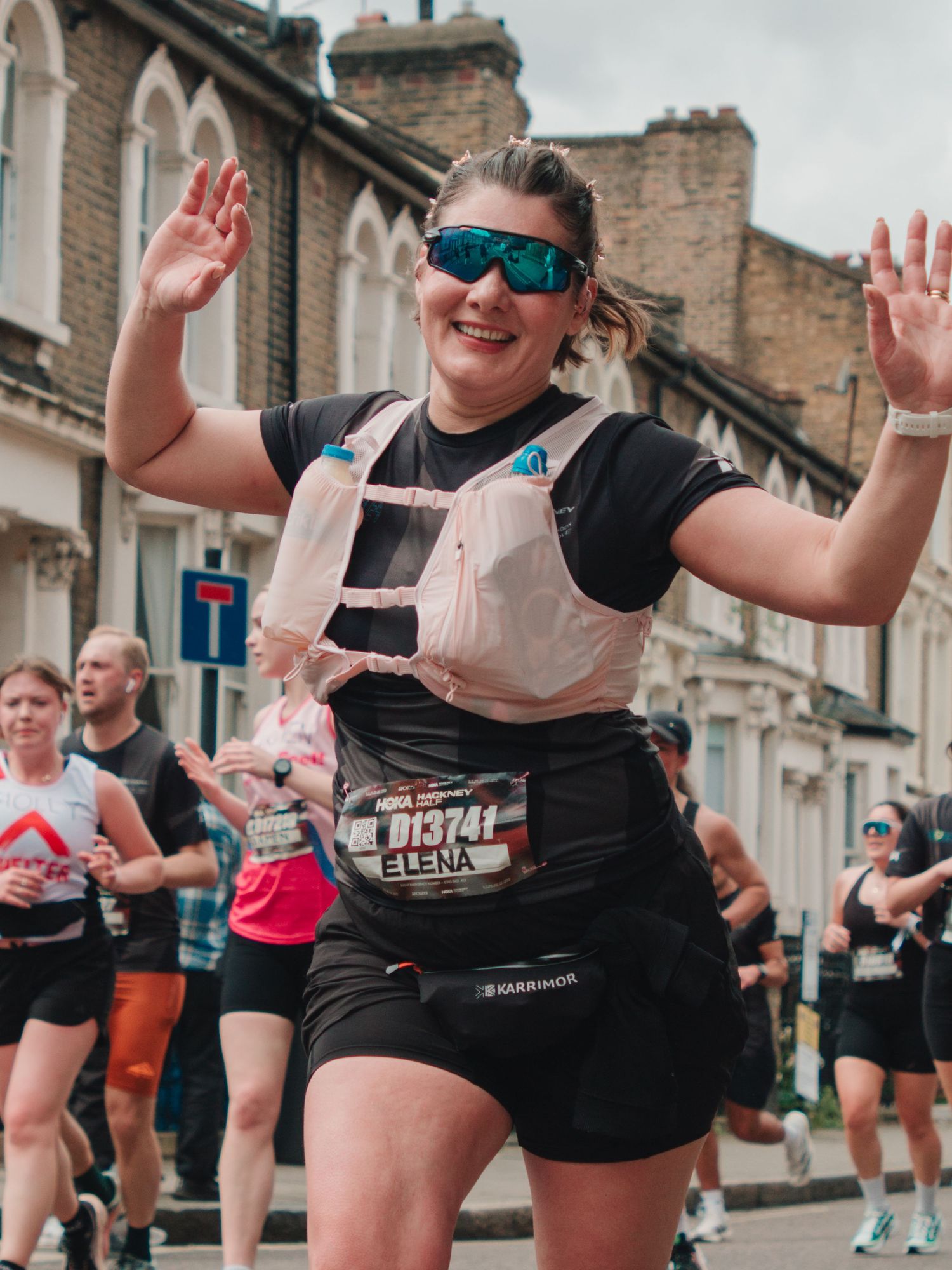 Runner named Elena wearing bib number 013741 and hydration vest celebrates during a street marathon race.