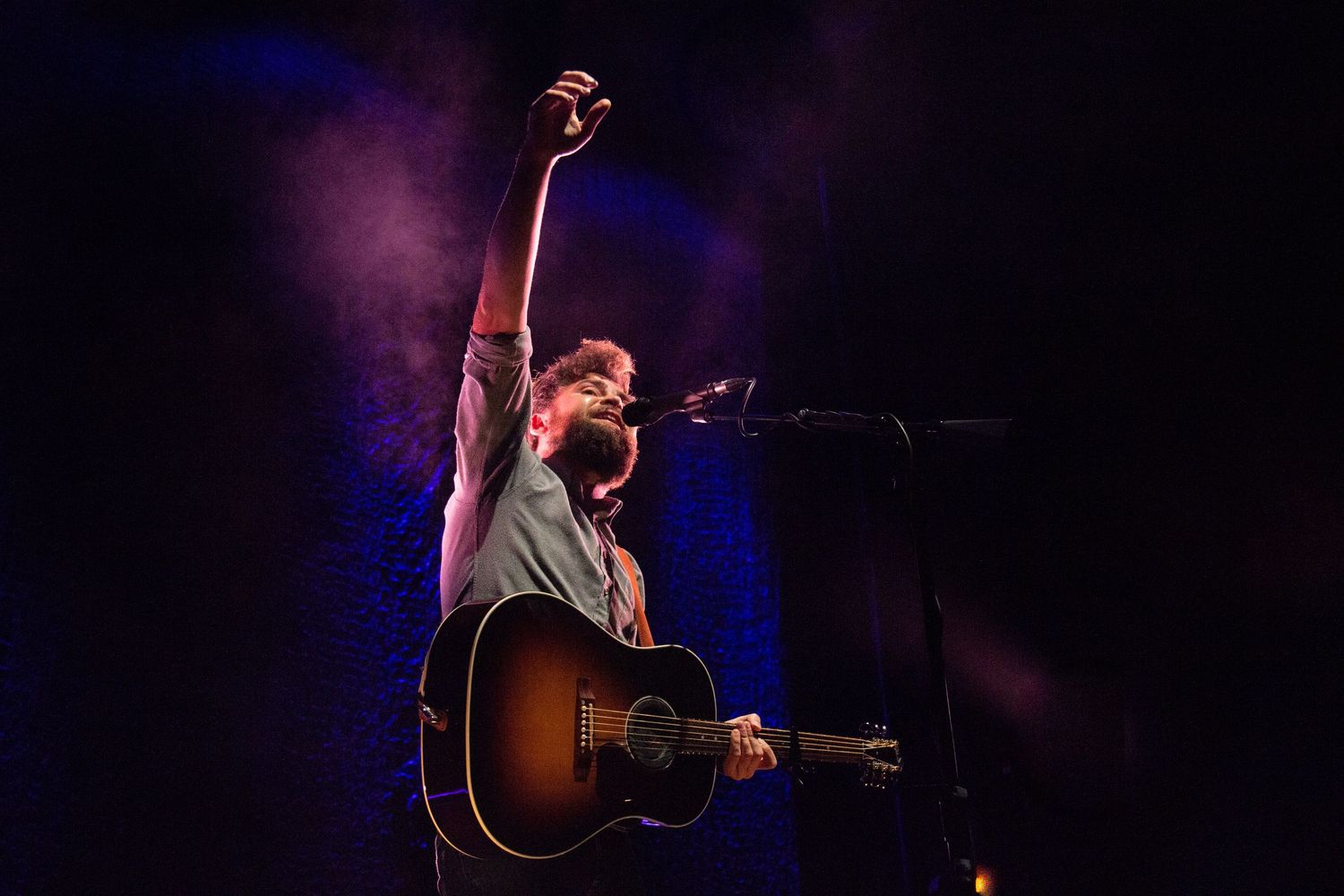 Concert performer plays acoustic guitar on dimly lit stage with dramatic blue lighting.