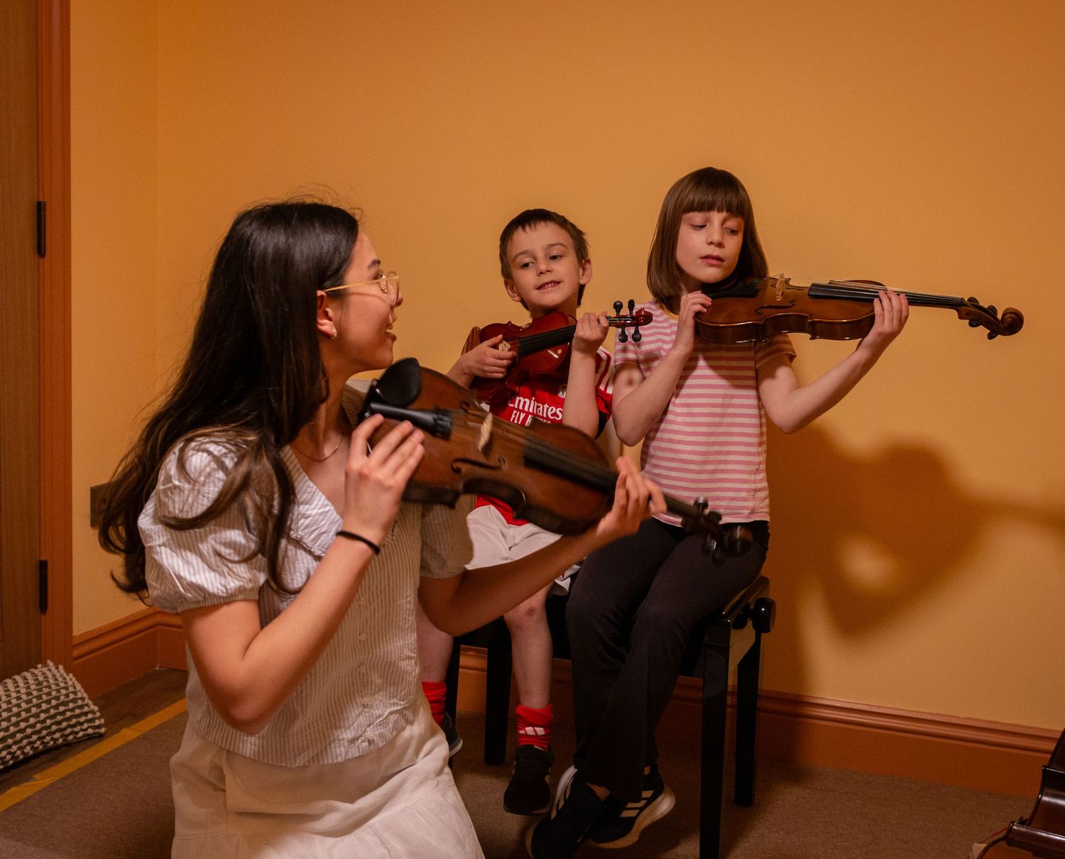 Three children playing violins together in a warm-lit room during a music lesson or practice session.