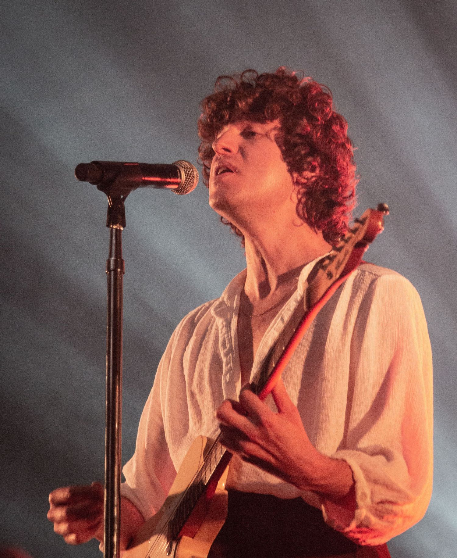 Performer in white shirt plays guitar and sings on smoky stage with dramatic lighting.