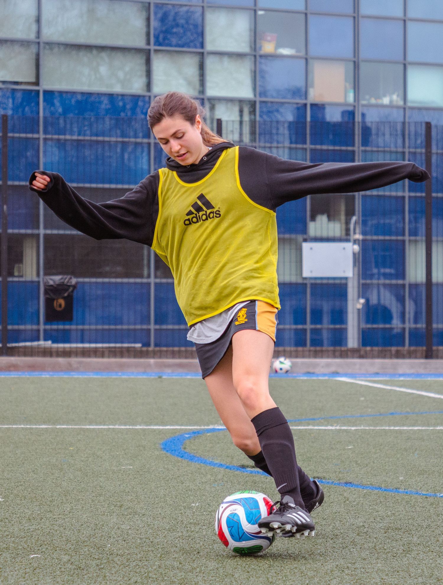 Soccer player in yellow training vest and black pants performs a kicking drill on artificial turf field.