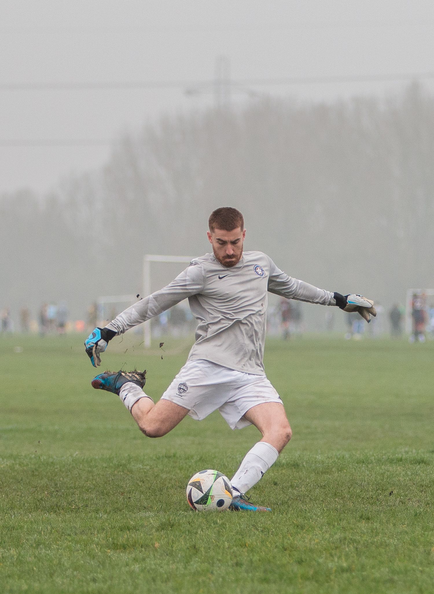 A soccer goalkeeper in white uniform makes a diving save during a misty match on a grass field.