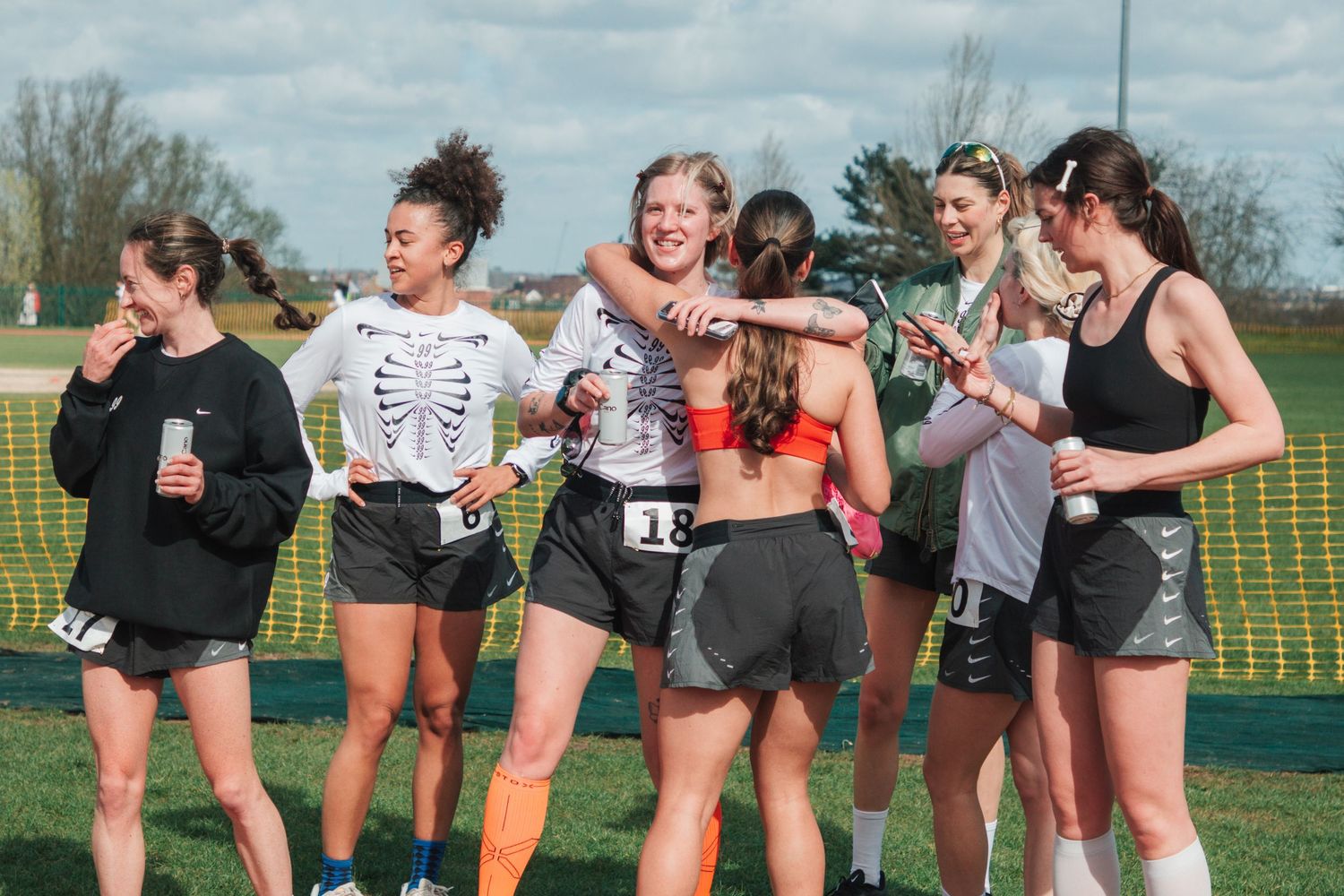 Female track athletes celebrate together on a green sports field on a sunny day, wearing race numbers and athletic wear.