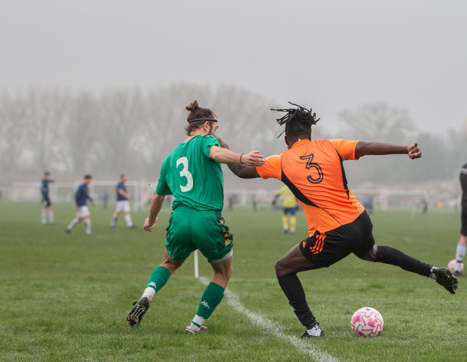 Soccer players in orange and green uniforms compete for the ball during a foggy match on a grass field.