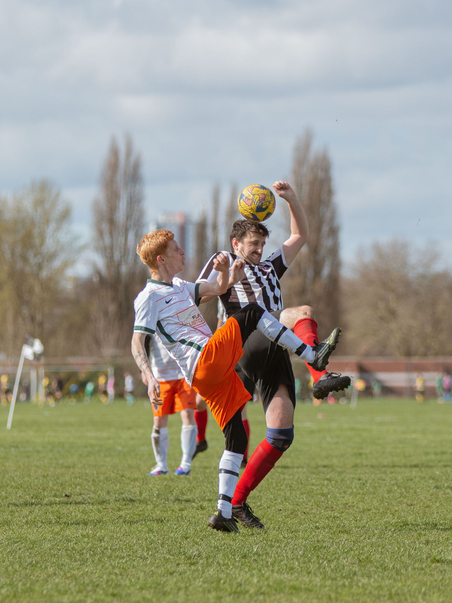 Soccer players collide in mid-air during an intense match on a grassy field with trees in the background.