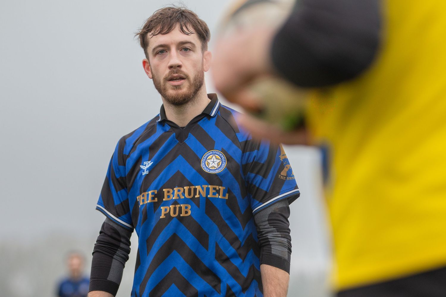 Soccer player wearing blue and black striped jersey makes a play during competitive match against yellow backdrop.