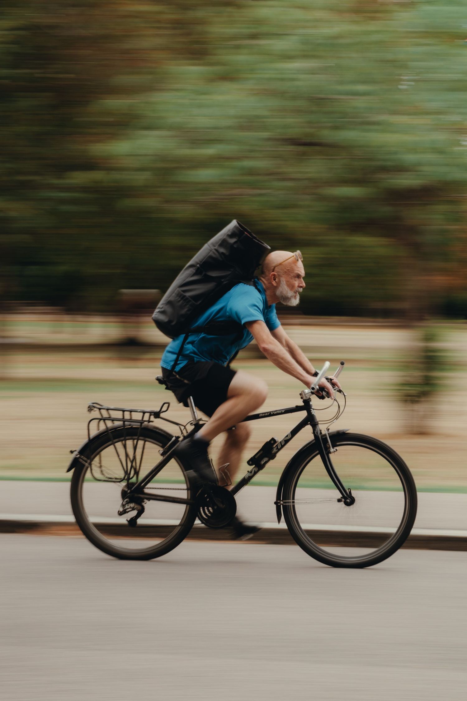Elderly man with white beard cycling fast on black bicycle, wearing blue t-shirt and backpack, motion blur background.