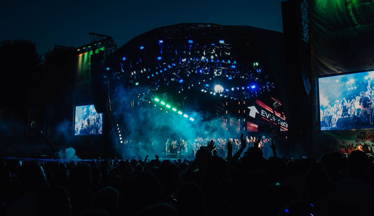 Panoramic view of outdoor concert stage with blue and green lighting effects illuminating the night sky.